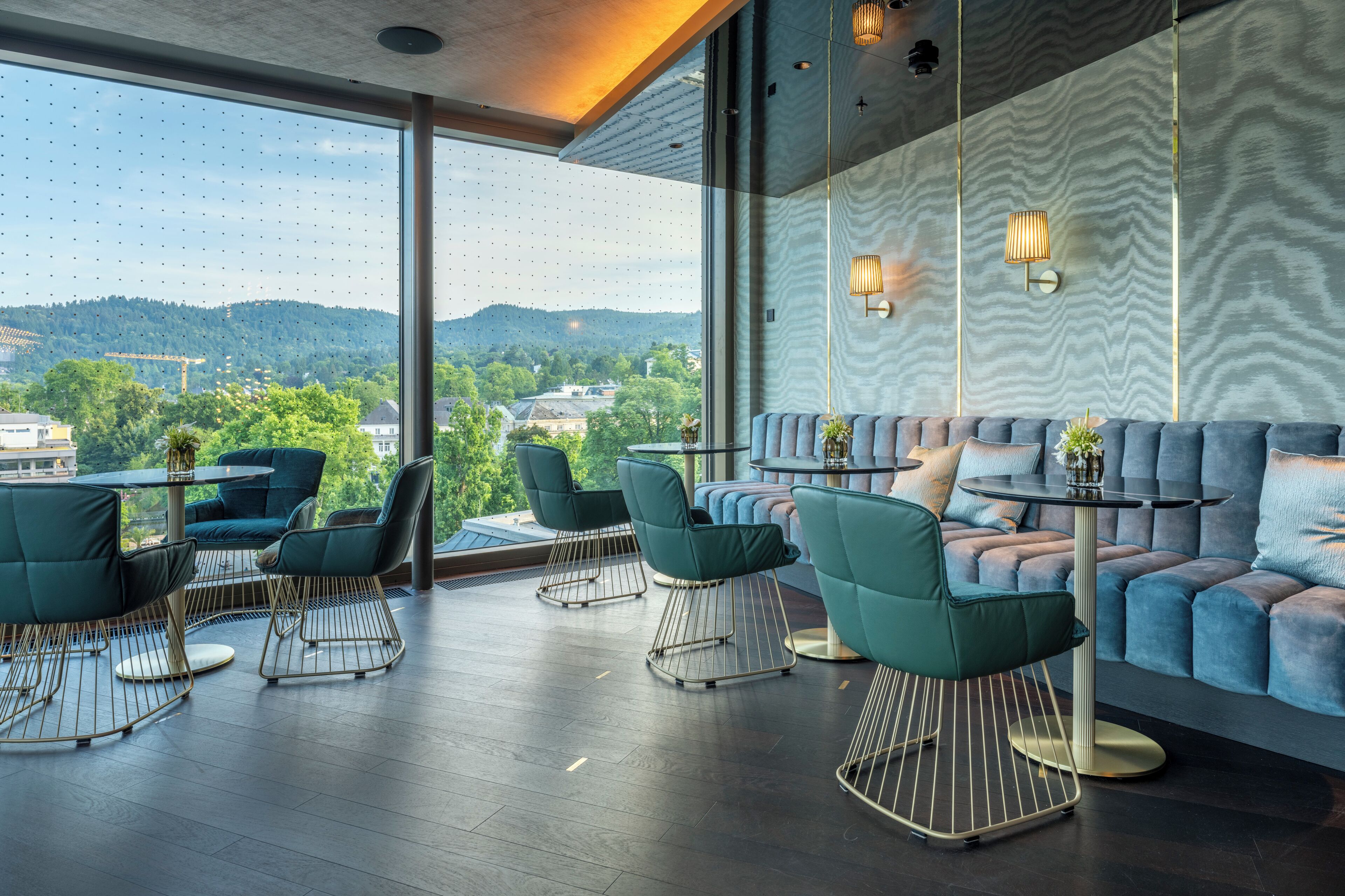 Ein moderner Loungebereich mit großem Fenster und Blick auf Stadt und Hügellandschaft. Mehrere grüne Polsterstühle und eine gepolsterte Couch, an deren Wand sich ein Muster befindet.