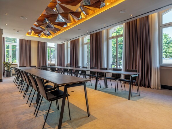 Une salle de conférence dotée d’une longue table et de chaises, de grandes fenêtres avec des rideaux et d’un plafond au design géométrique.