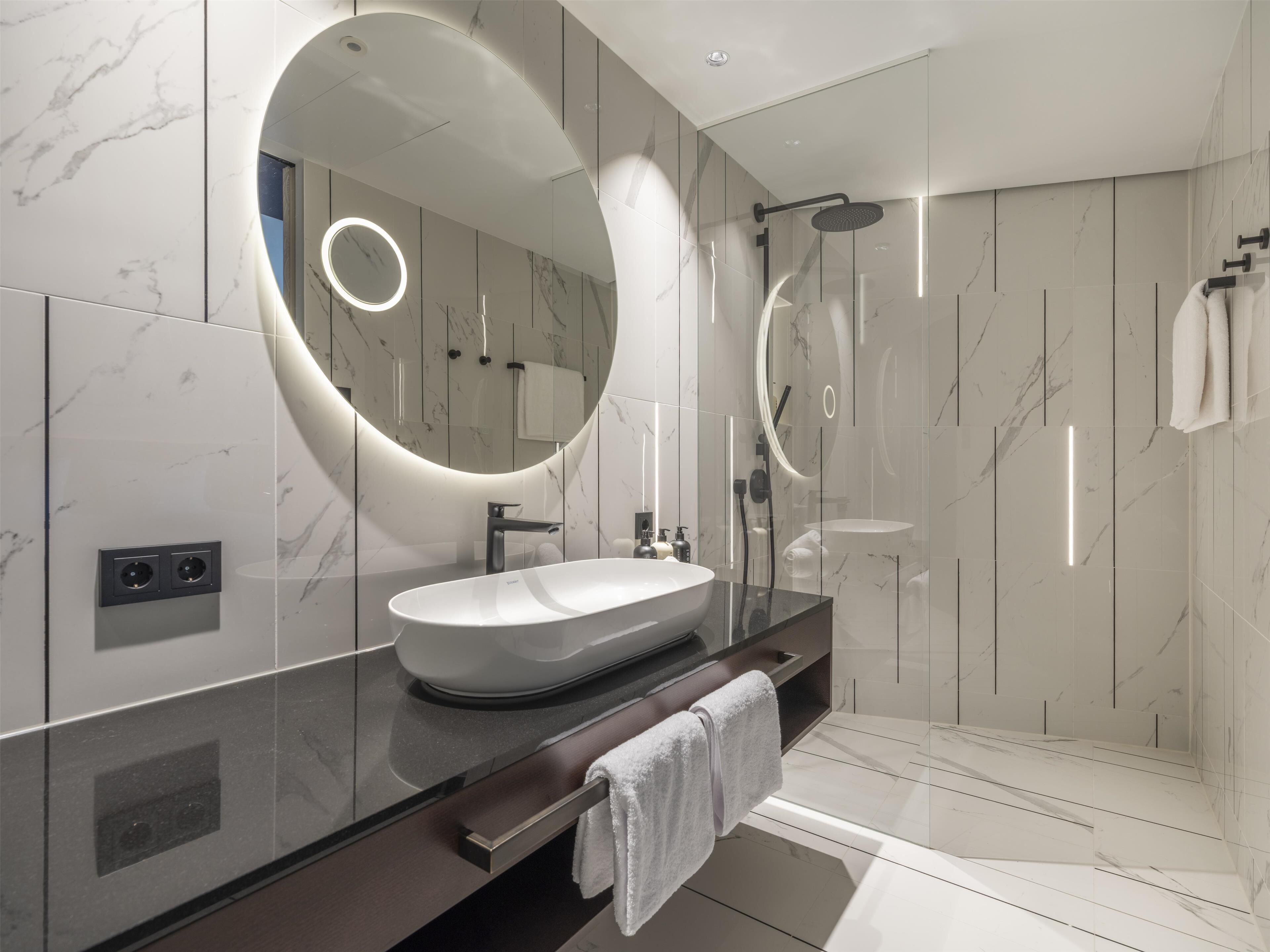 A modern bathroom with a large oval mirror, a vessel sink, and a glass-enclosed shower.