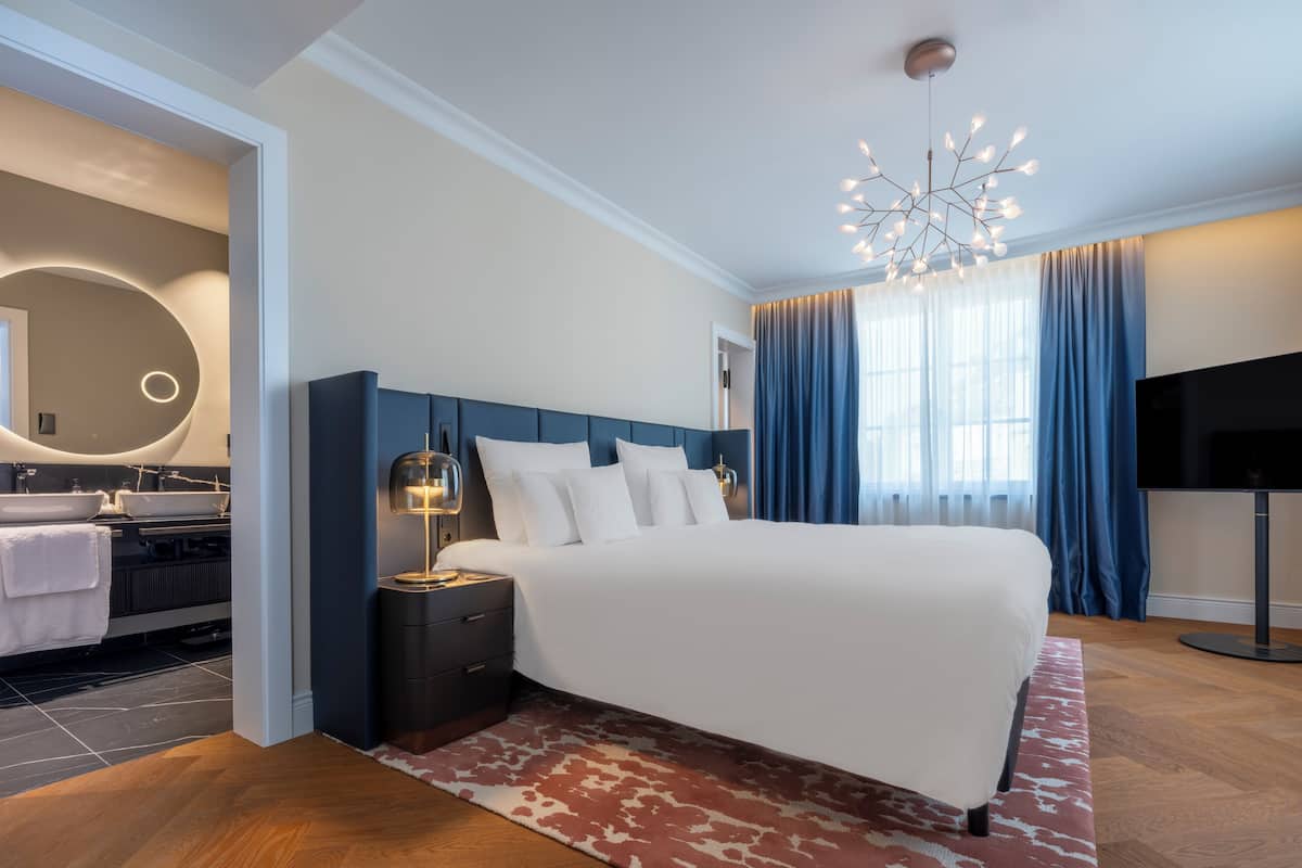 Une chambre d’hôtel moderne dotée d’un grand lit, d’une table de chevet avec une lampe, d’une télévision et d’une salle de bain avec un grand miroir rond et deux lavabos.