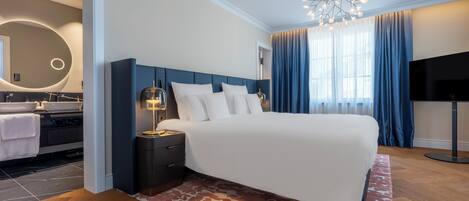 Una habitación de hotel moderna con una cama grande, una mesita de noche con lámpara, un televisor y un baño con un espejo redondo y dos lavamanos.