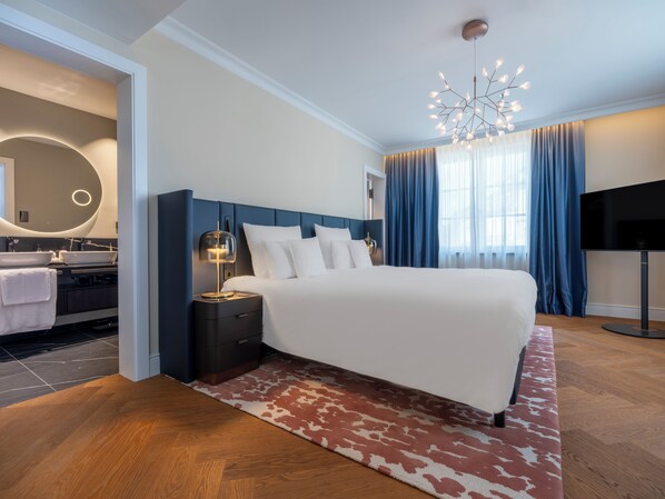 Una habitación de hotel moderna con una cama grande, una mesita de noche con lámpara, un televisor y un baño con un espejo redondo y dos lavamanos.