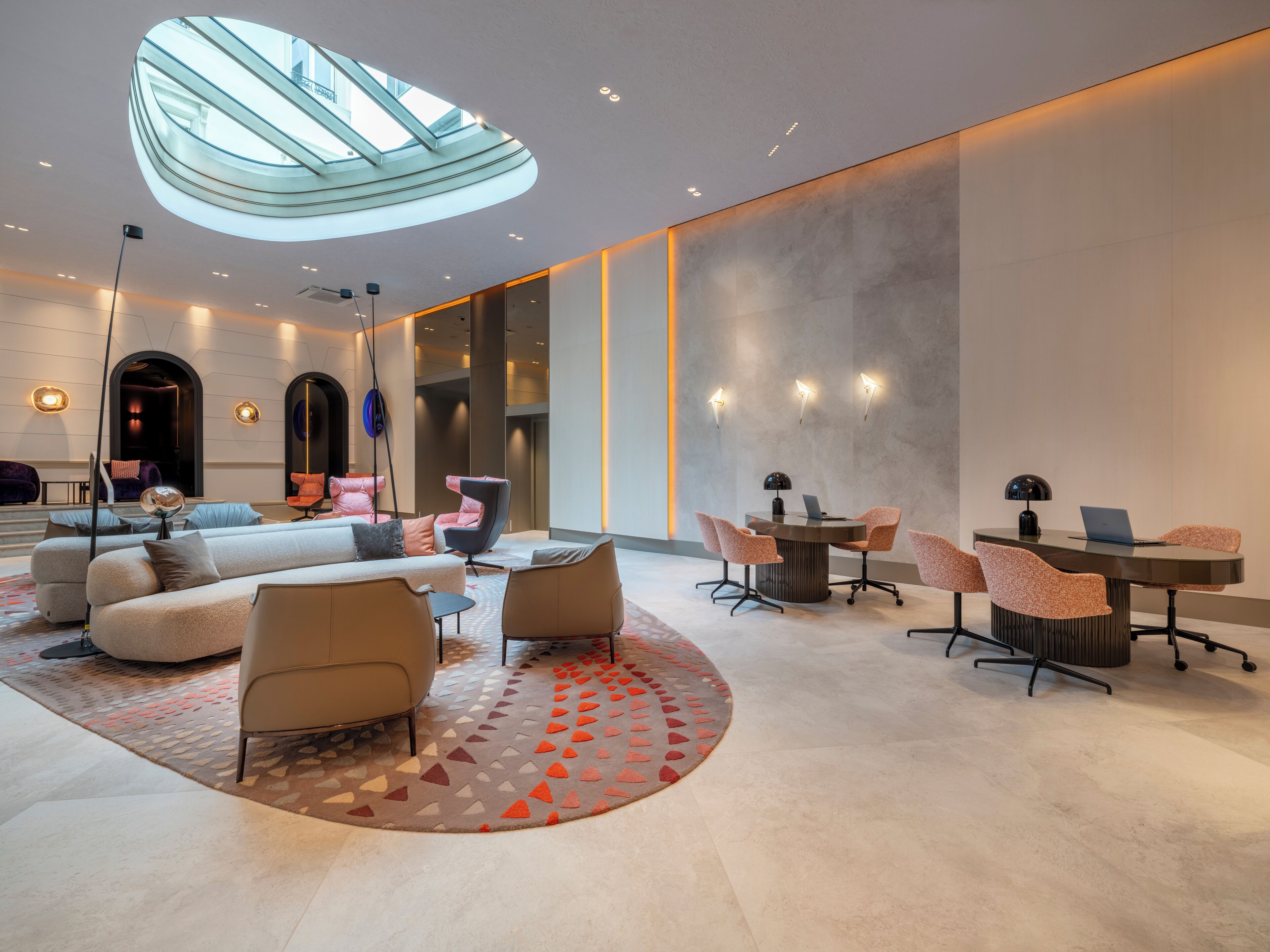 A modern hotel lobby with a central seating area, a round table with chairs, and a glass ceiling.