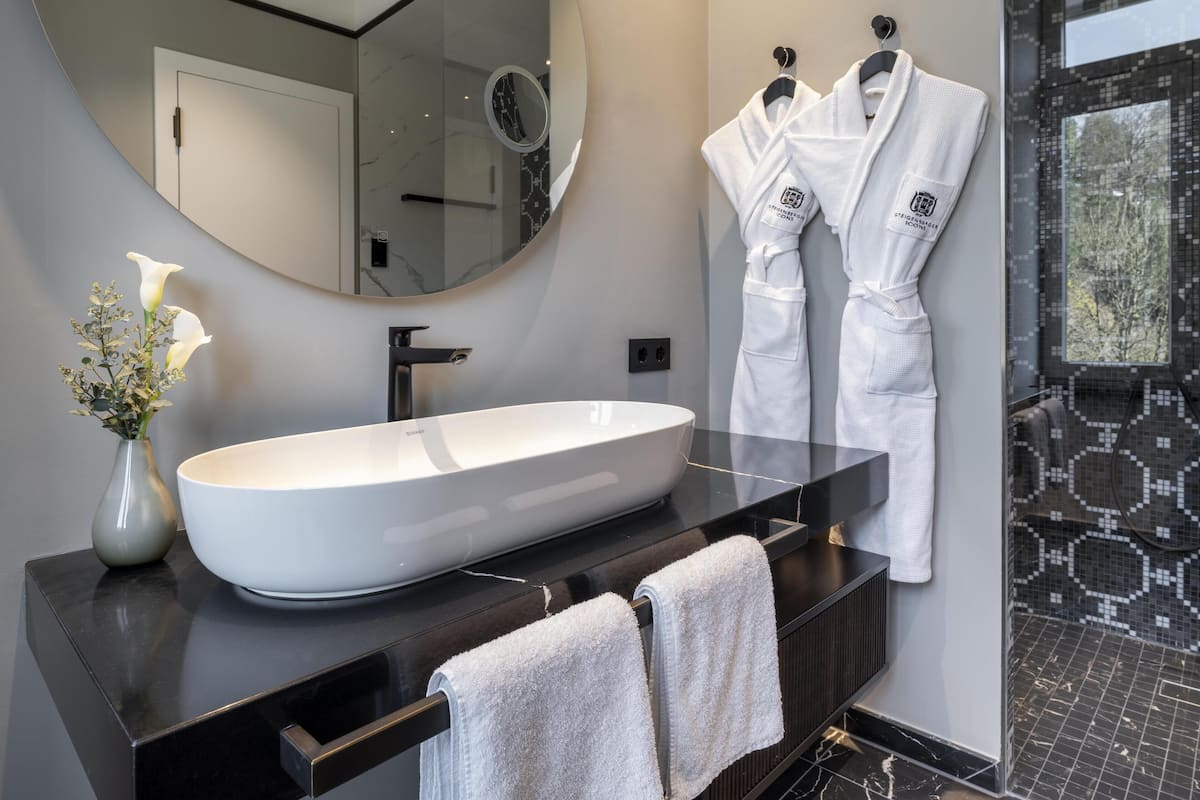 Une salle de bain moderne avec un grand lavabo ovale, un miroir rond et deux peignoirs blancs suspendus à des crochets.