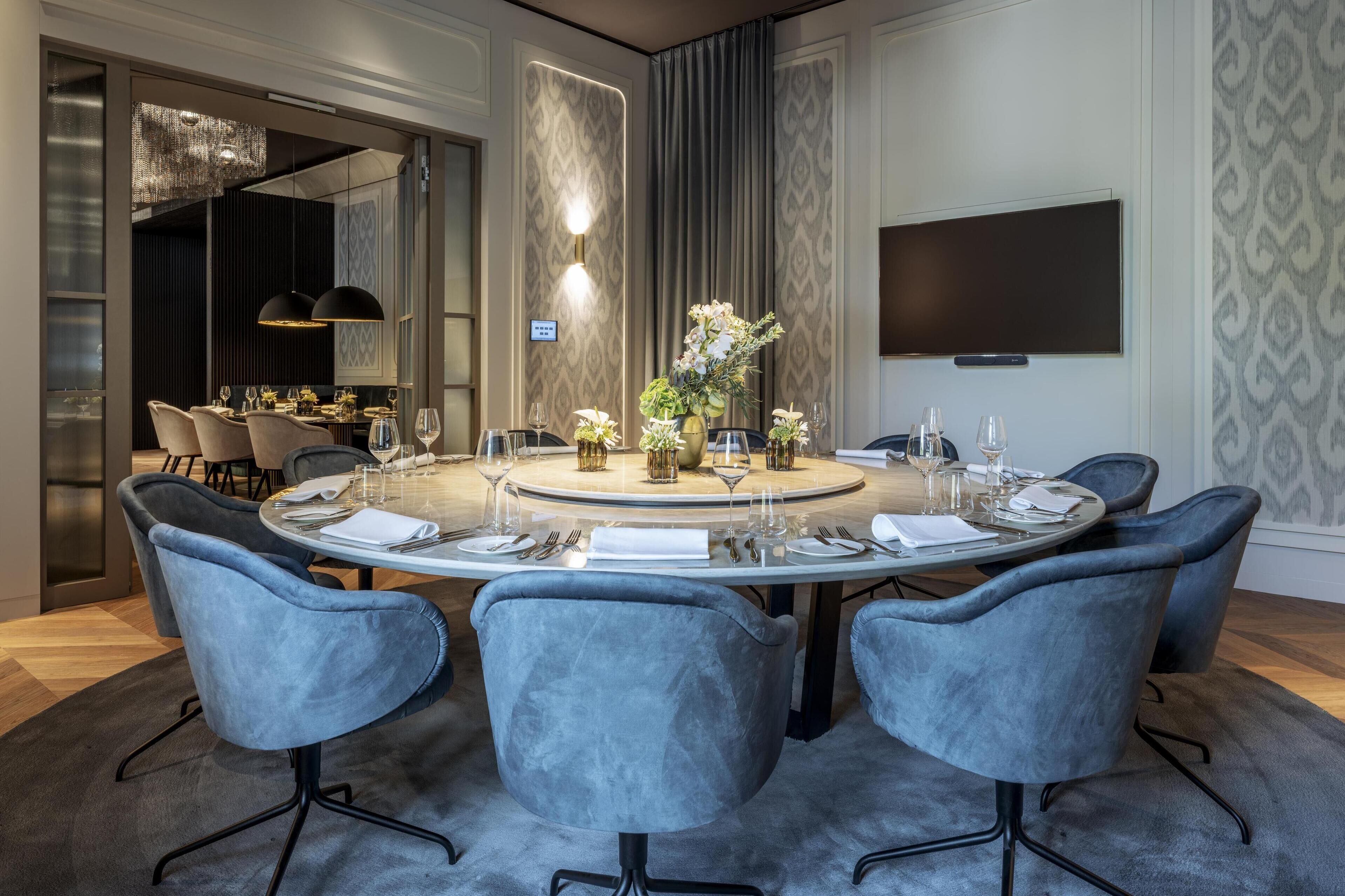 Un coin repas moderne avec une table ronde dressée pour six personnes, entourée de six chaises grises à revêtement rembourré. Un vase rempli de fleurs est placé au centre de la table.
