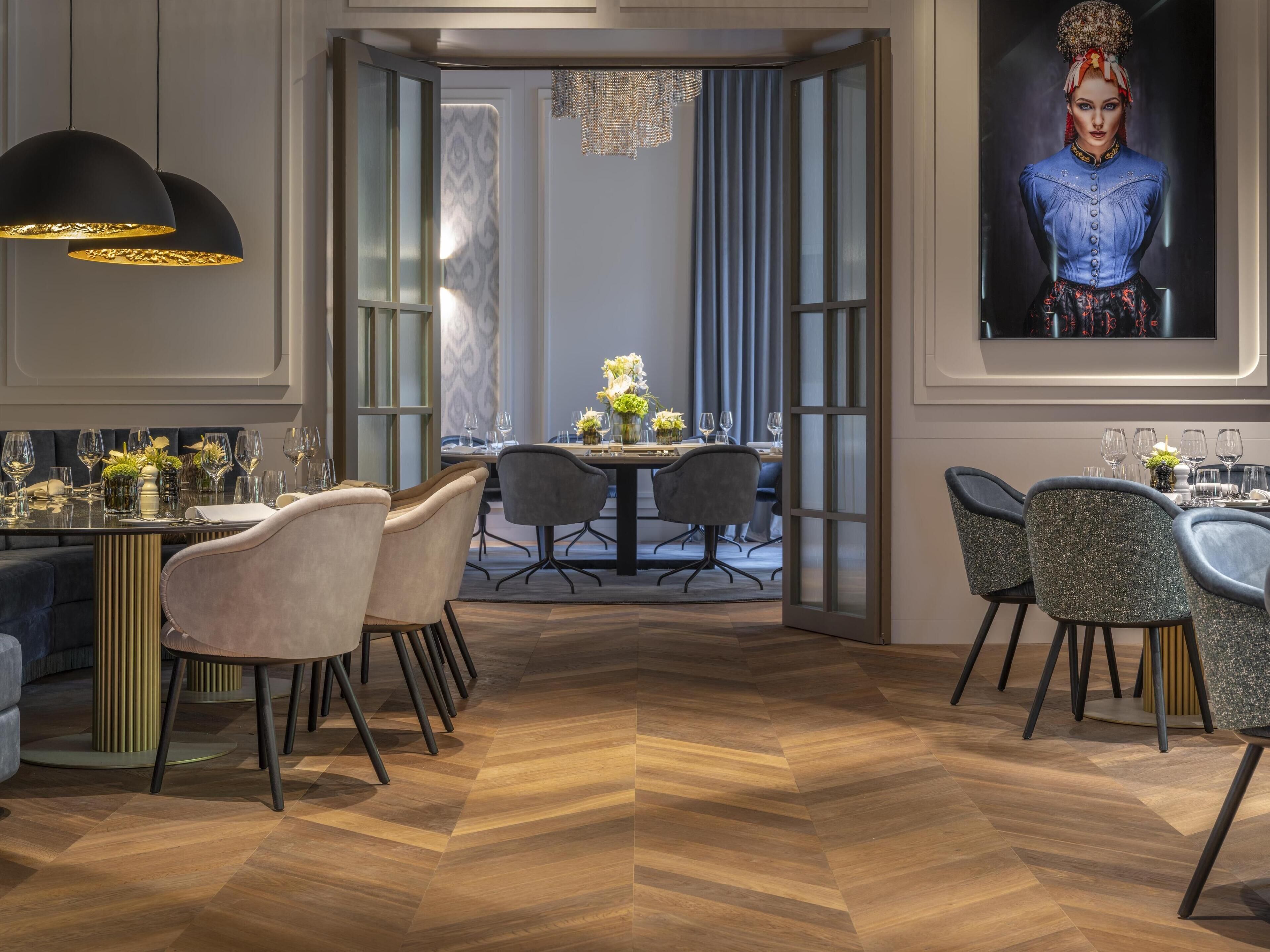 A modern dining area with a wooden herringbone floor, a central round table with chairs, and a chandelier.