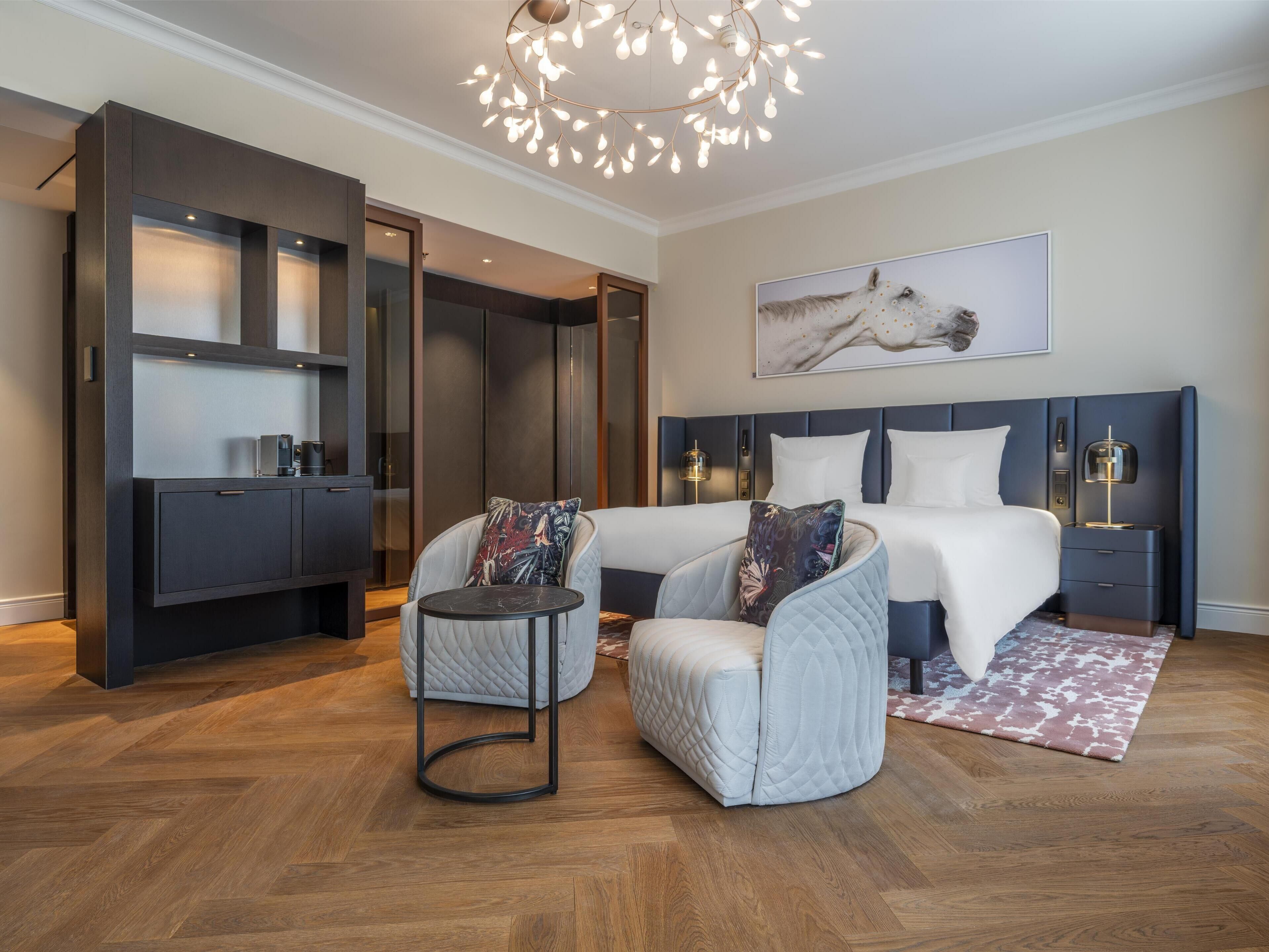 A modern hotel room with a large bed, a seating area with two chairs, a desk, and a wardrobe.