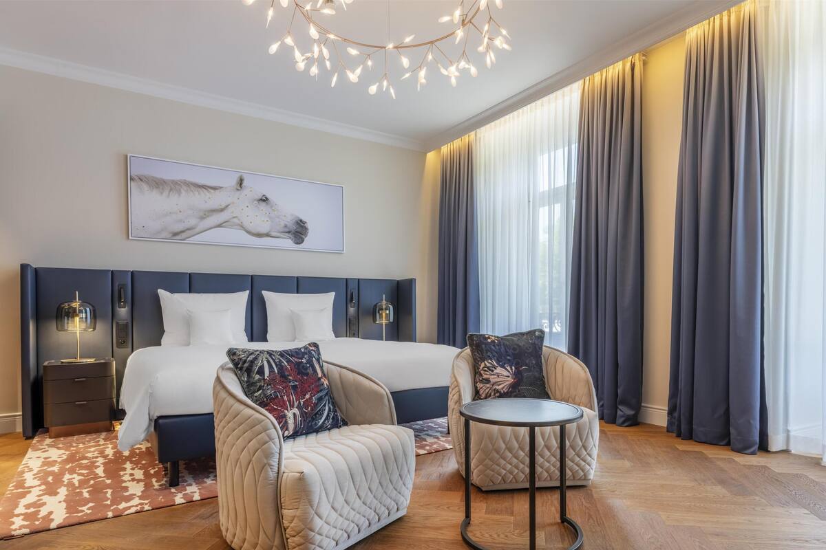 Une chambre d’hôtel moderne avec un grand lit, deux fauteuils, une petite table ronde, une table de chevet, une lampe fixée au mur et un tableau représentant un cheval.