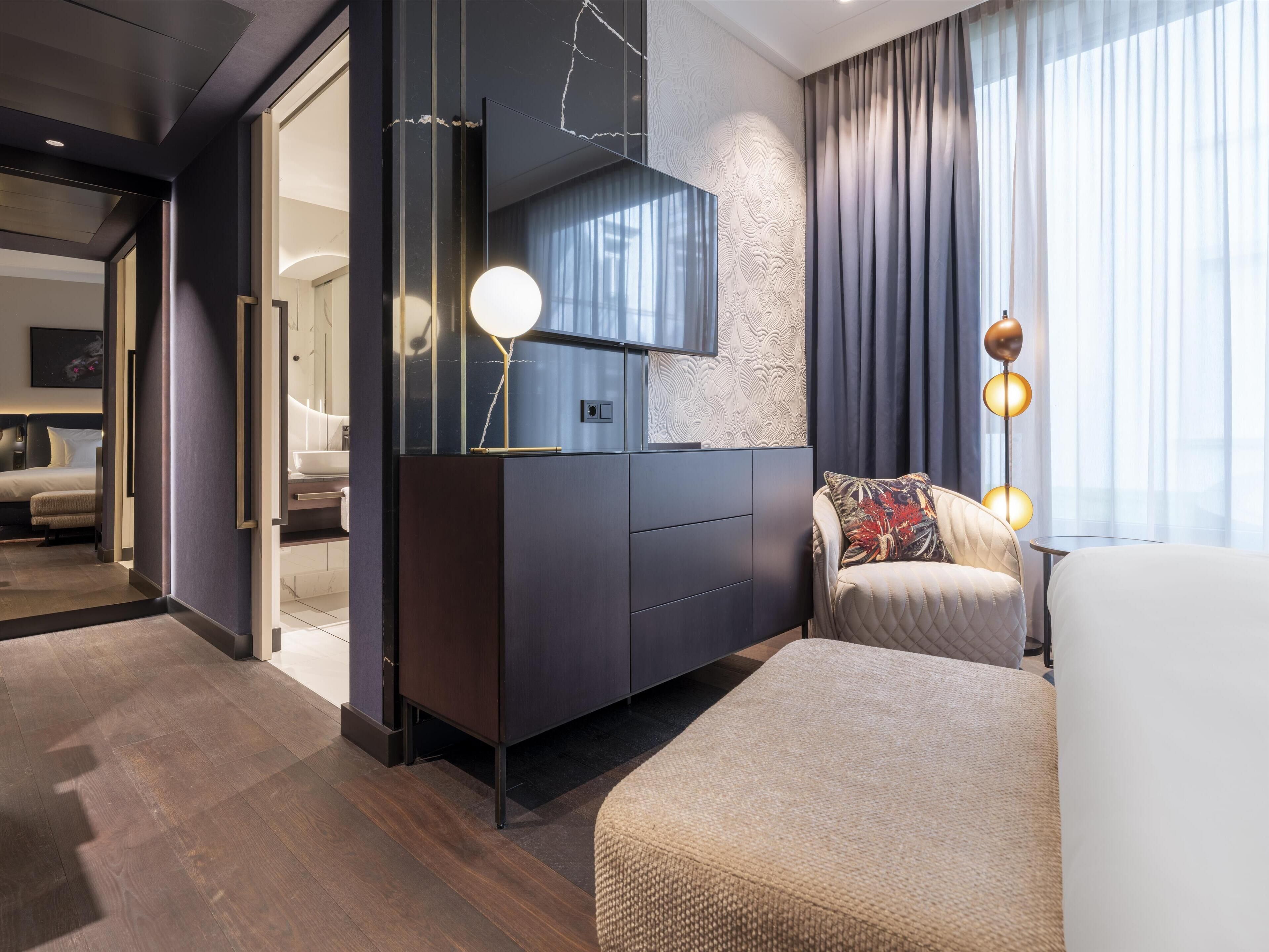 Una habitación de hotel moderna con piso de madera oscura, un televisor de pantalla plana grande montado en una pared con textura, una zona de estar acogedora con un banco acolchado y un ventanal con cortinas.