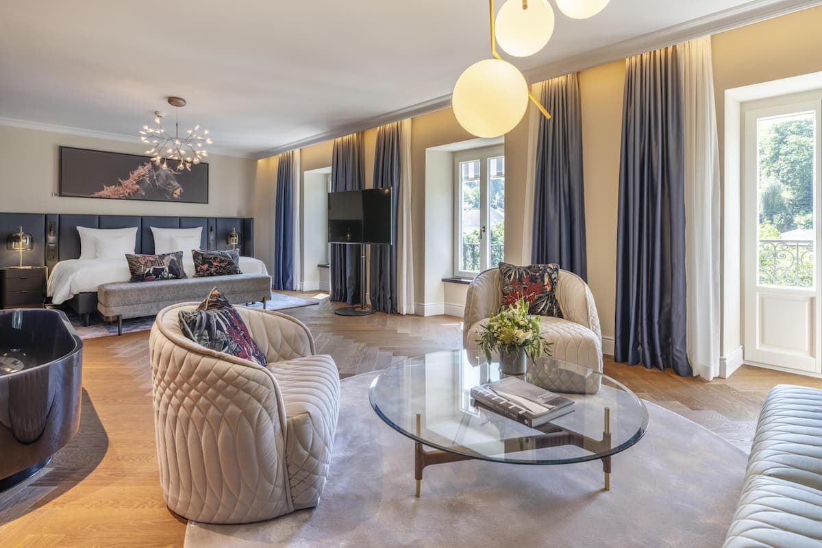 Un salon moderne avec un canapé, un fauteuil et une table basse en verre. On y trouve un lit avec une tête de lit et un téléviseur dans un coin.