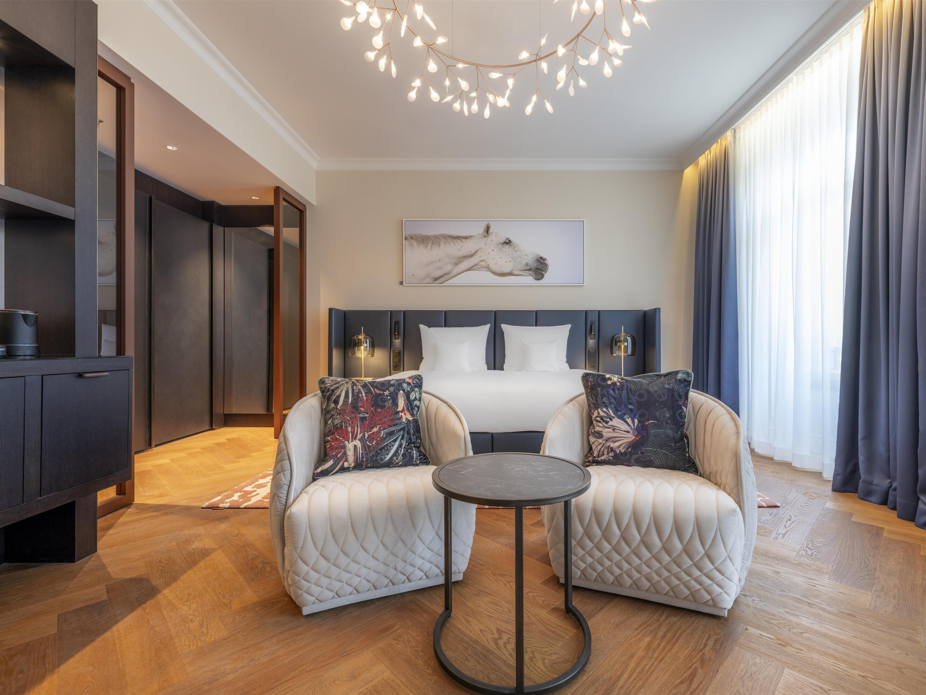 Una habitación de hotel moderna con una cama, dos sillones, una mesita, una lámpara de techo y un cuadro en la pared.