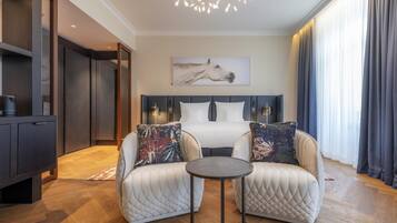 Una habitación de hotel moderna con una cama, dos sillones, una mesita, una lámpara de techo y un cuadro en la pared.