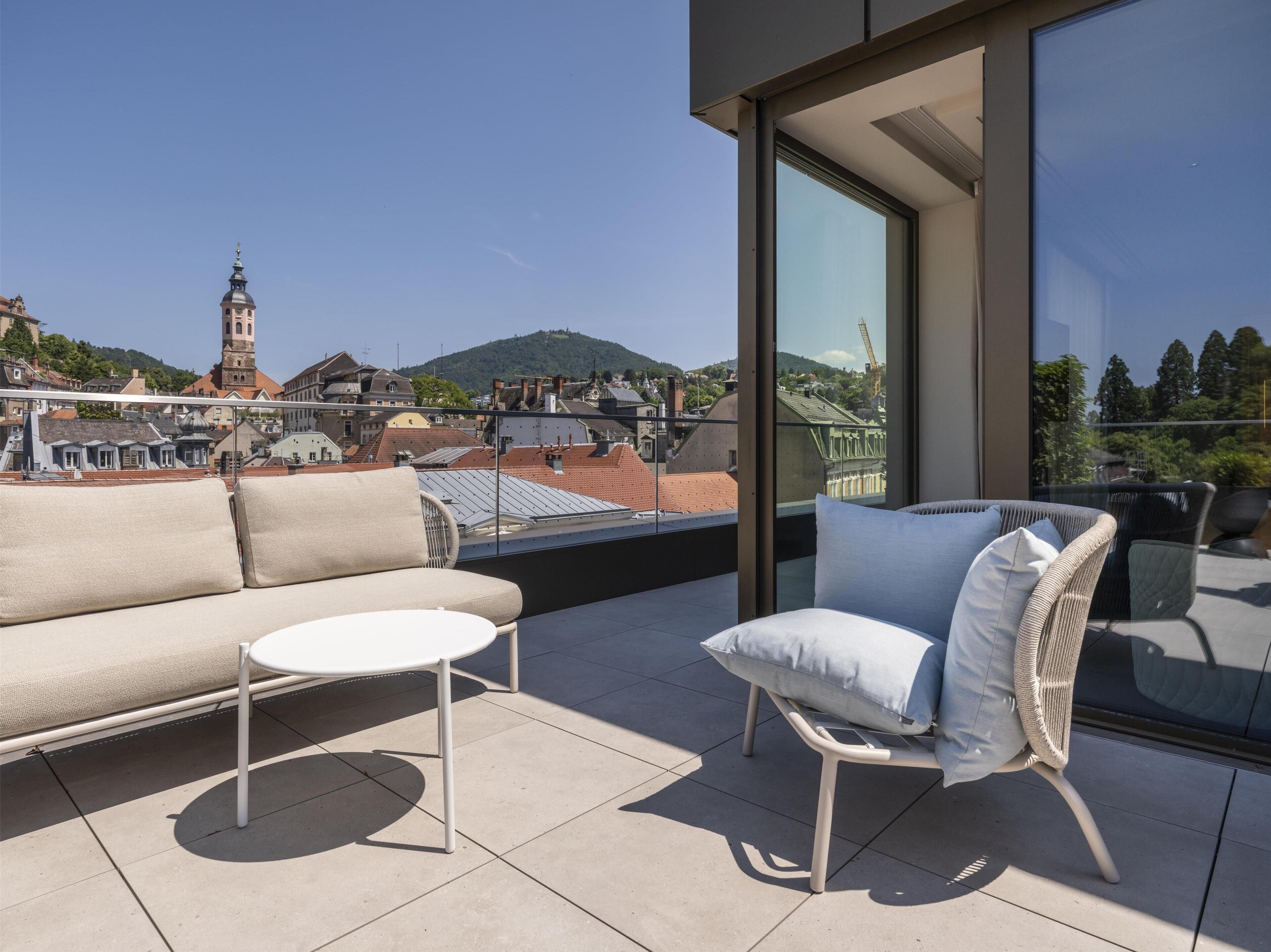 Un balcon avec un canapé et une chaise, donnant sur un paysage urbain avec des immeubles et une colline.