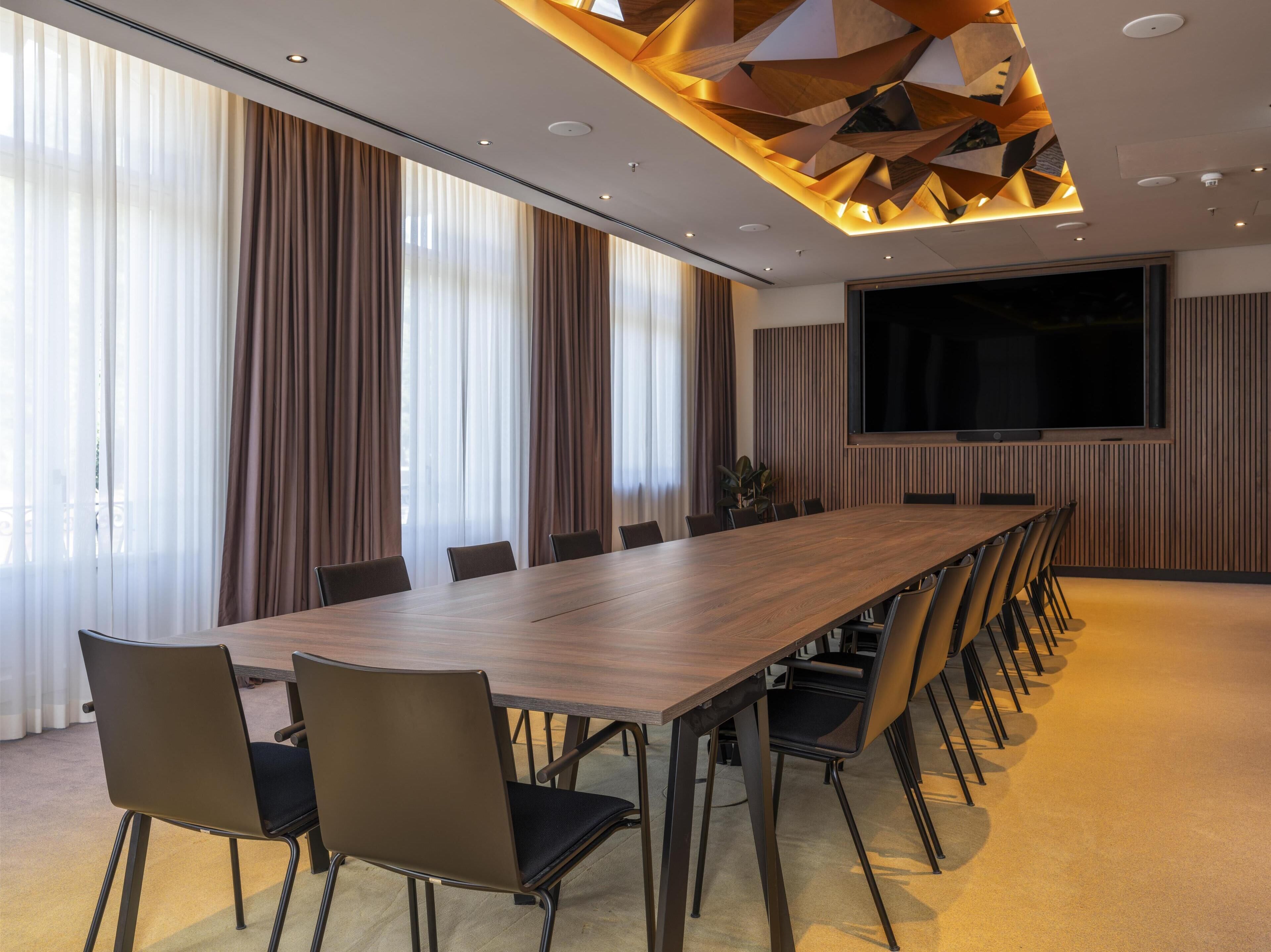 Uma sala de conferências com uma mesa comprida, cadeiras, uma tela grande e um design moderno no teto.