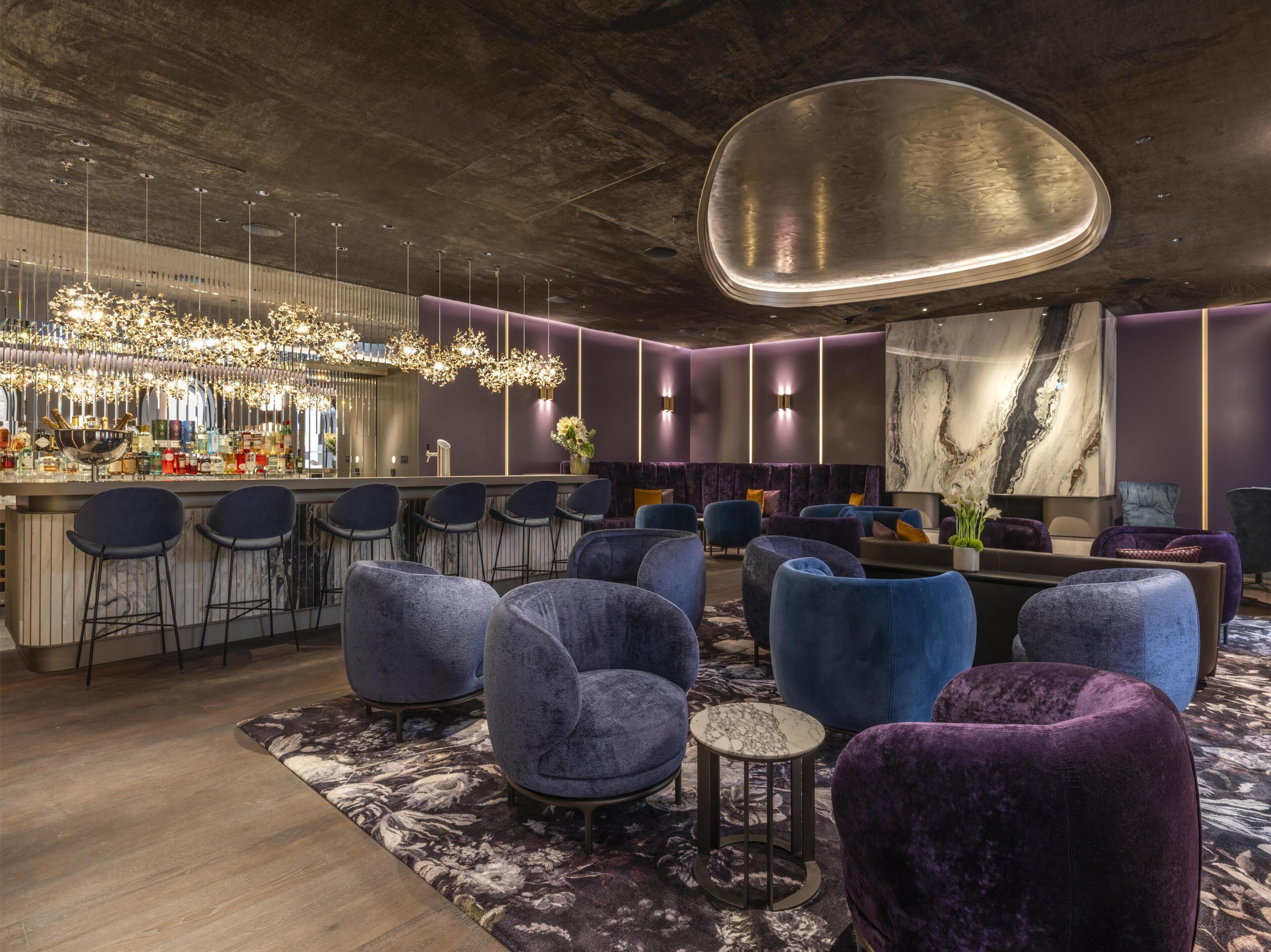 A modern bar area with a bar counter, multiple seating options including bar stools and cushioned chairs, and a decorative ceiling light fixture.