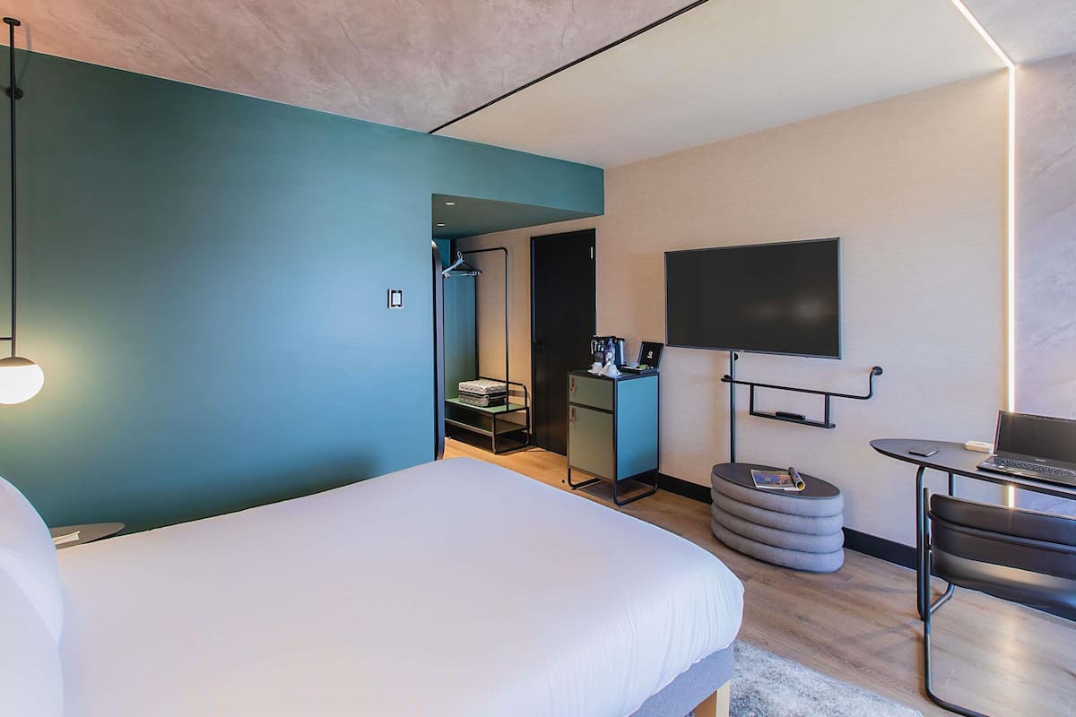 Ein modernes Hotelzimmer mit Bett, Schreibtisch mit Laptop, Wand-Fernseher und einer Garderobe.