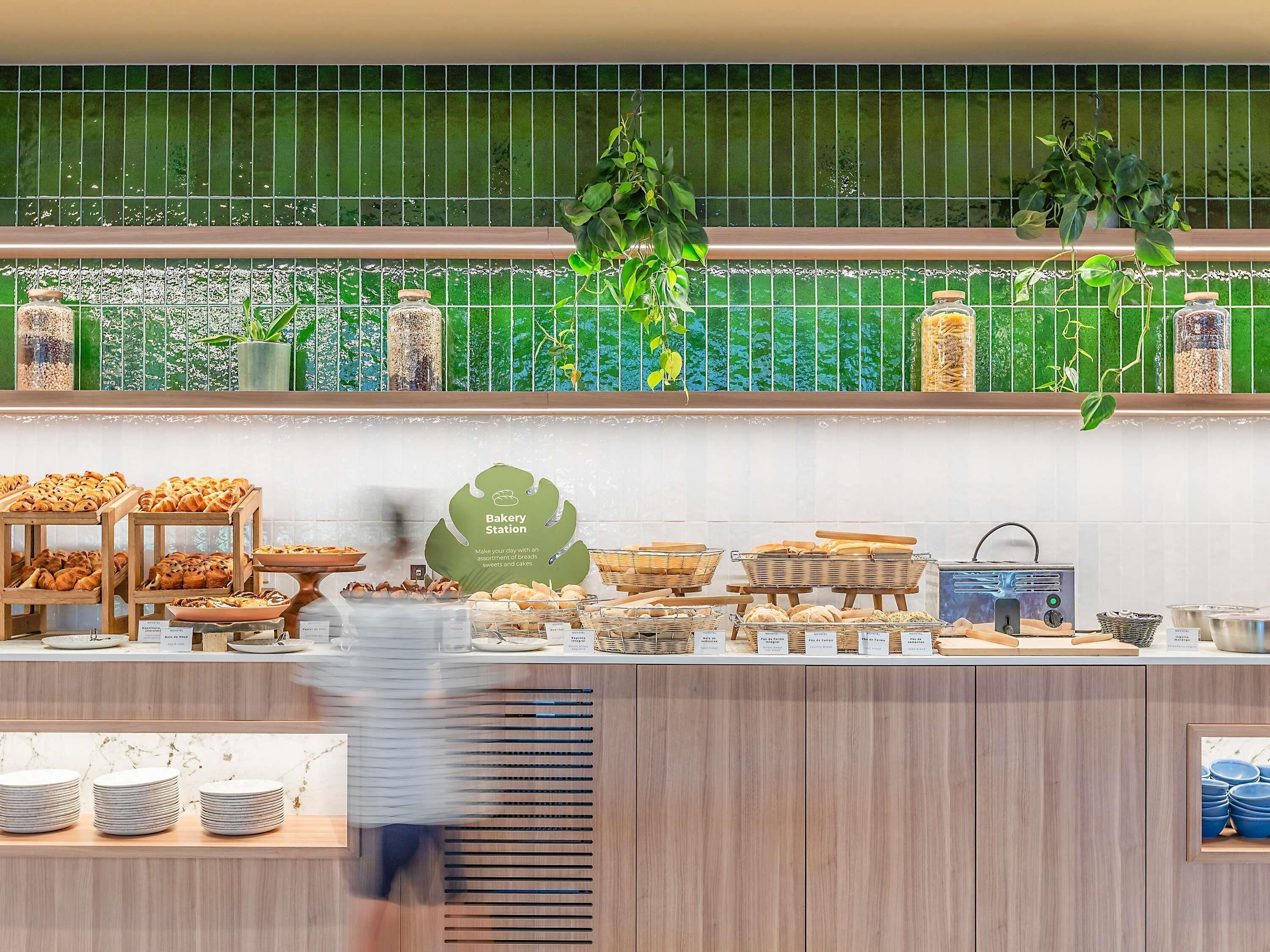 A bakery counter with various baked goods, a sign reading 'Bakery Station,' and a tiled backsplash with green tiles and hanging plants.