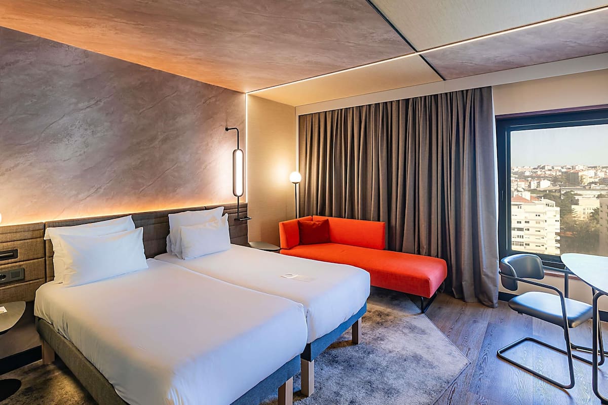 Ein Hotelzimmer mit Bett, einer roten Couch, einem Schreibtisch mit Stuhl und Blick auf die Stadt durch das Fenster.