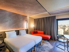 Habitación de hotel con cama, sofá rojo, escritorio con silla y vista a la ciudad por la ventana.
