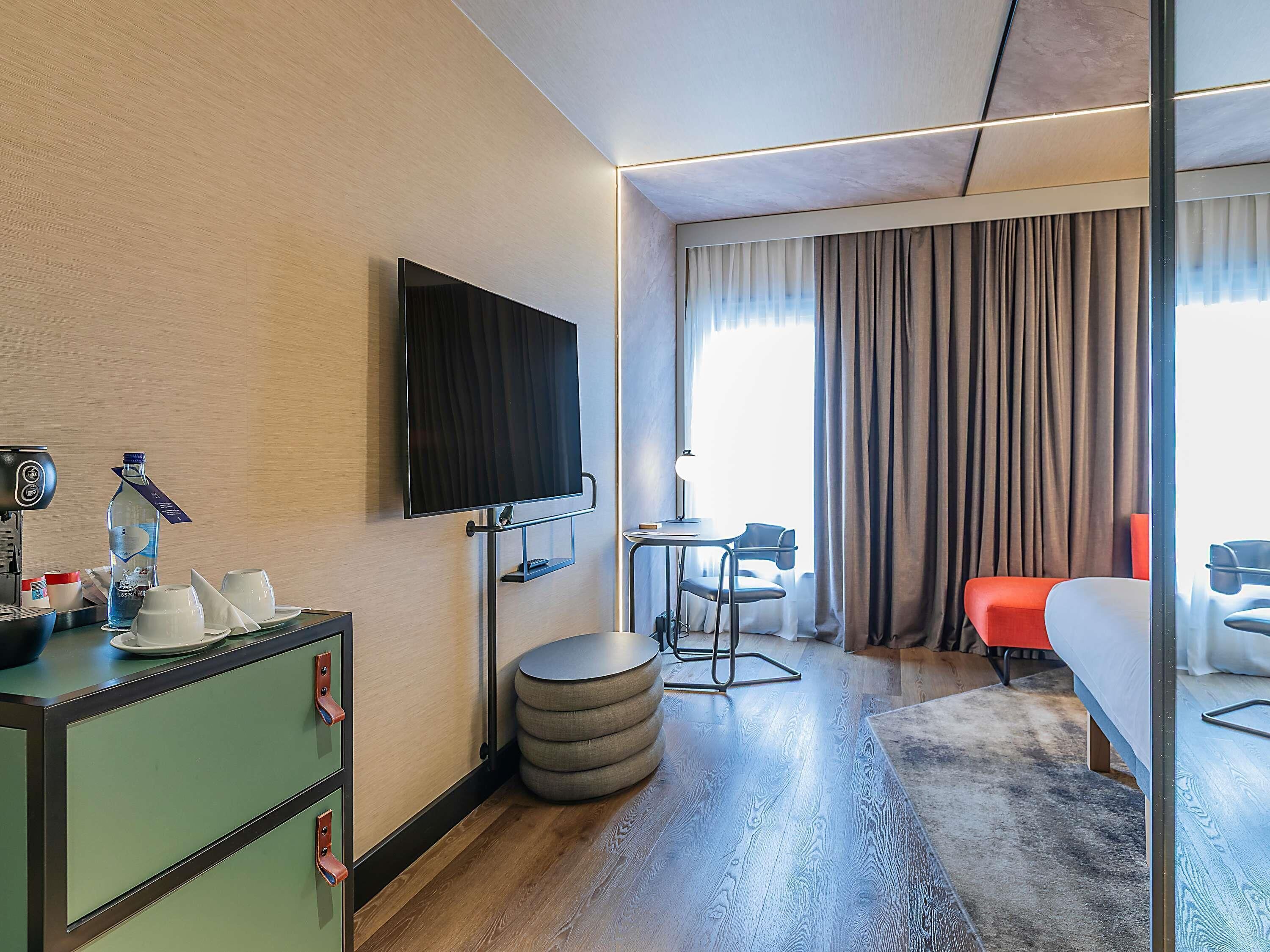 Une chambre d’hôtel équipée d’un téléviseur à écran plat, d’un bureau avec une lampe, d’une chaise, d’un lit et d’une grande fenêtre avec des rideaux.