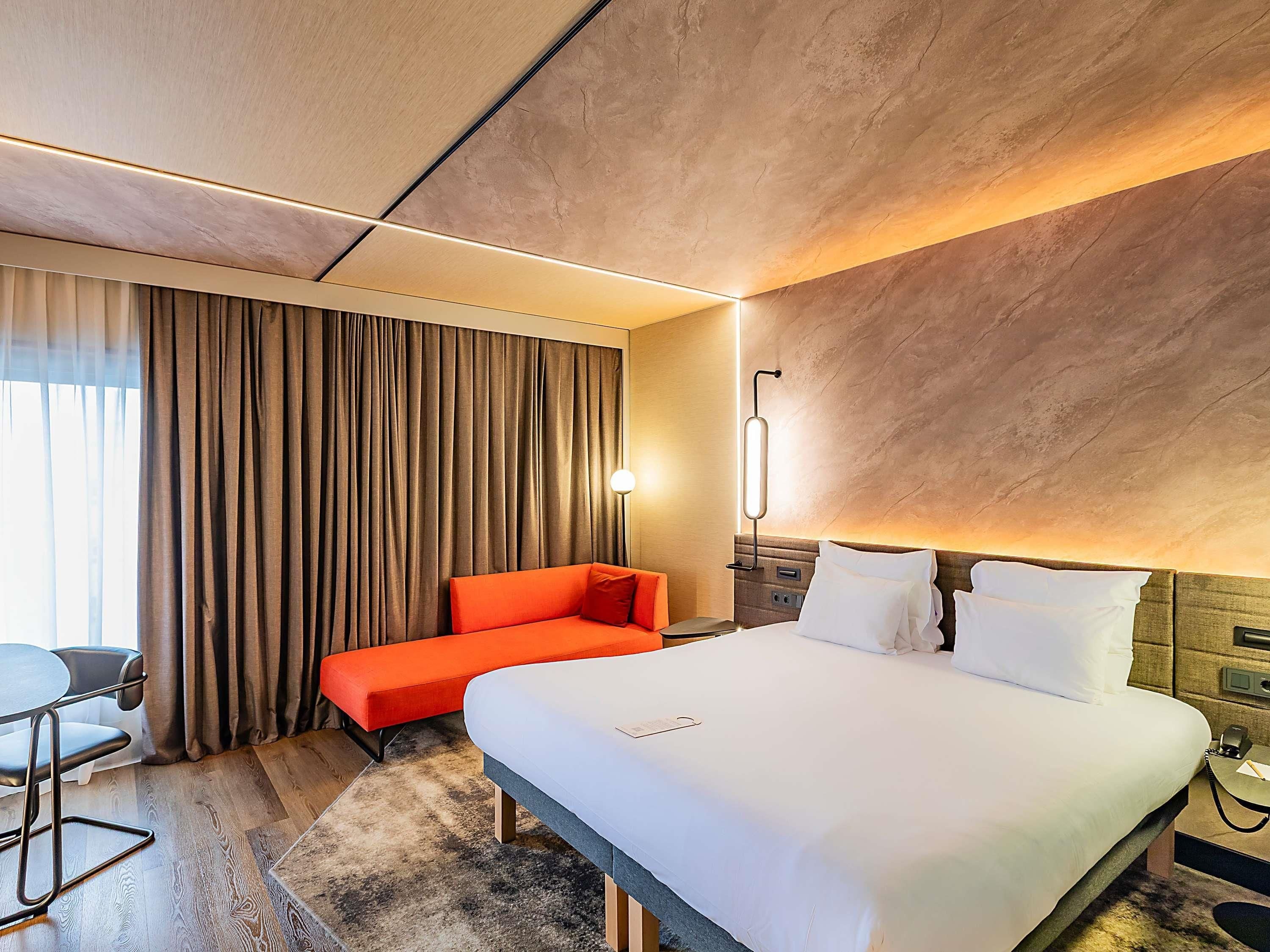 A hotel room with a bed, a red sofa, a desk, and a large window with curtains.