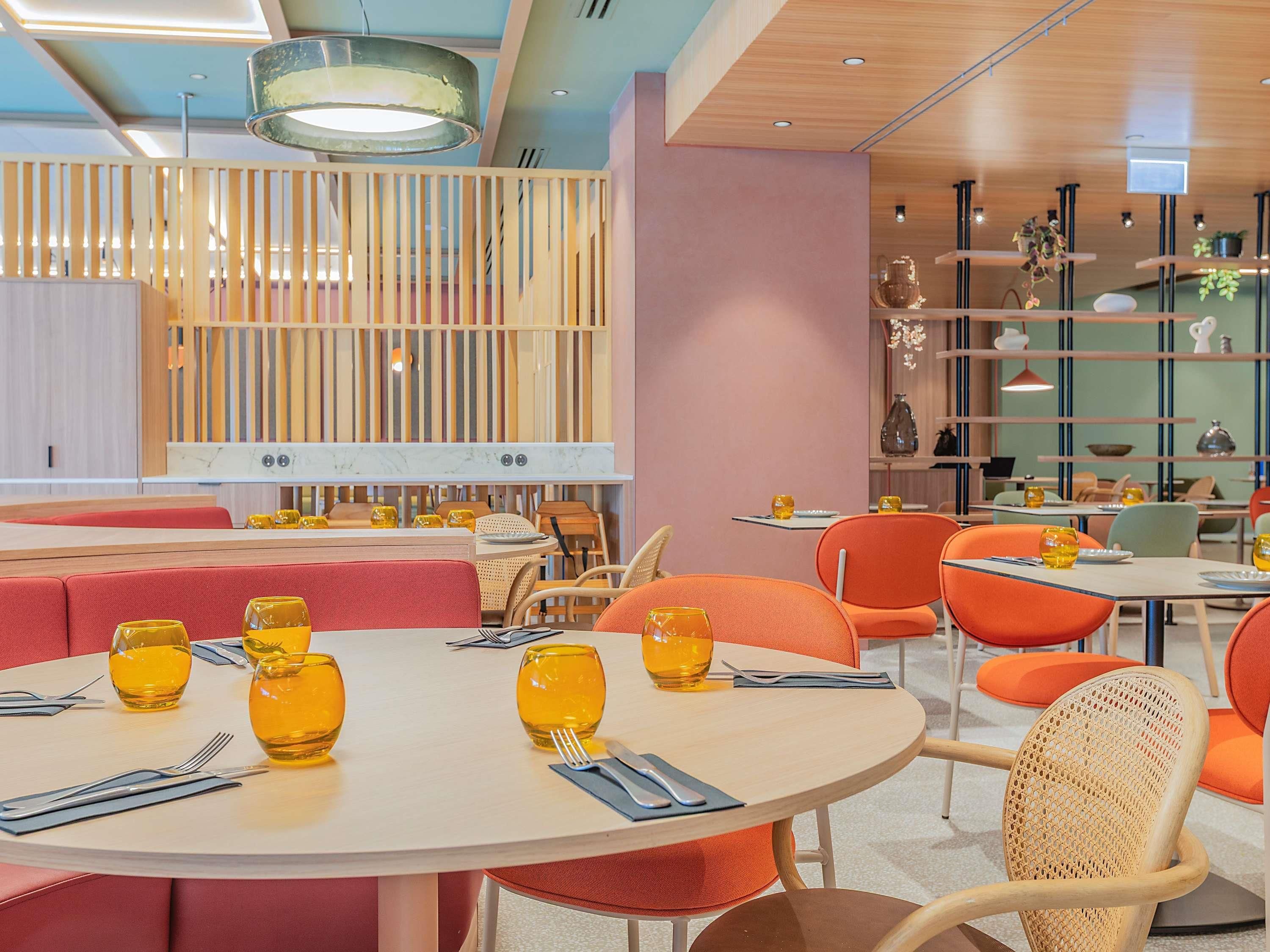 Een moderne eetruimte met ronde tafels, oranje stoelen en geel glaswerk.
