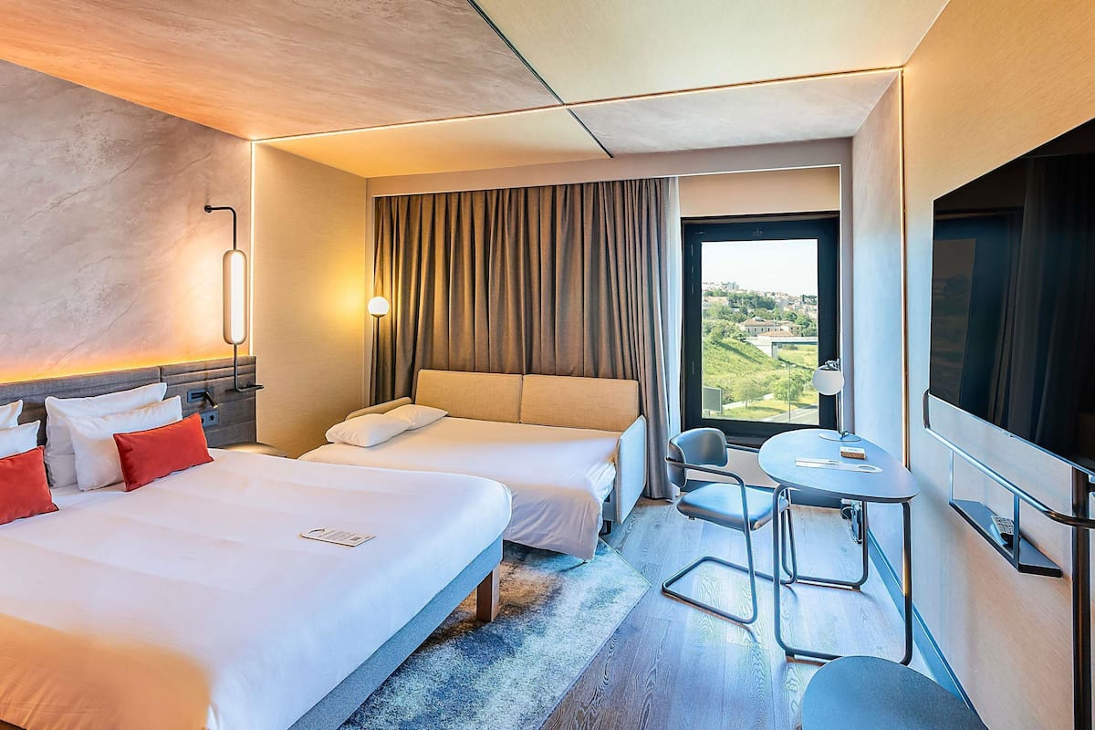 Ein Hotelzimmer mit zwei Betten, einem Schreibtisch, einem Stuhl, einem großen Fenster mit Aussicht und einem Flachbildfernseher.