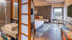 A hotel room with a bathroom, a bed, a desk, and a view of the city. - Novotel Lisboa (Lisbon)