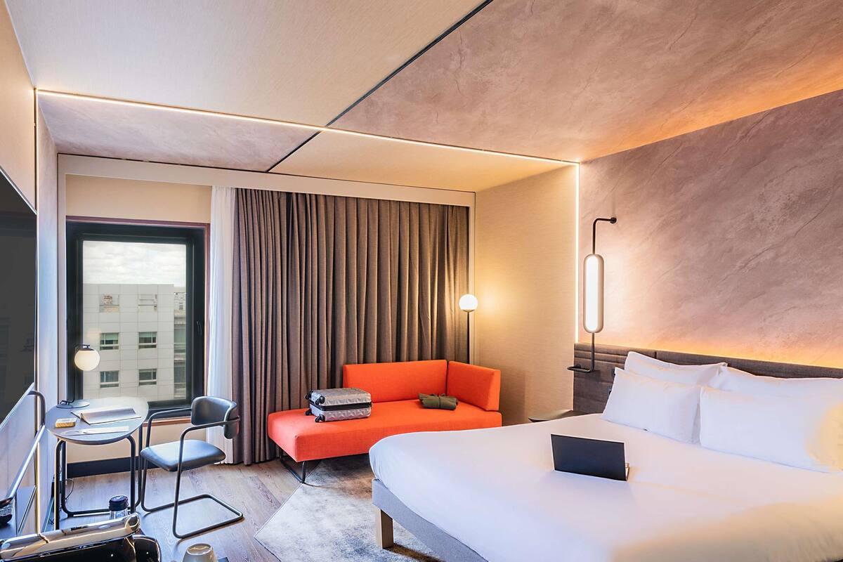 Una habitación de hotel con una cama, un escritorio con una silla, un sofá naranja pequeño y una ventana con cortinas.