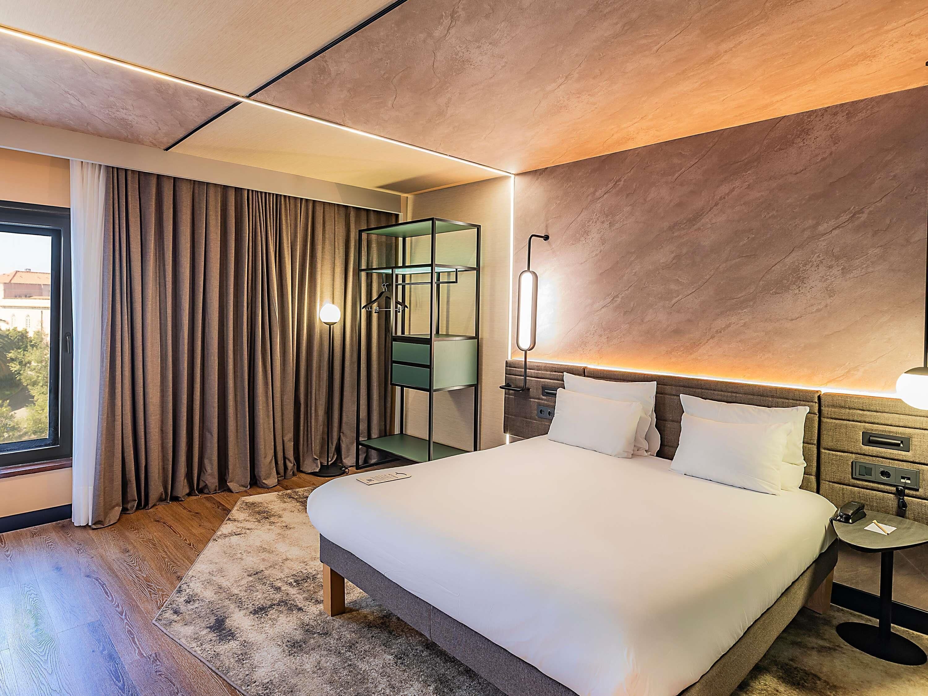 A hotel room with a large bed, a bedside table with a lamp, a wardrobe, and a view of the outdoors.