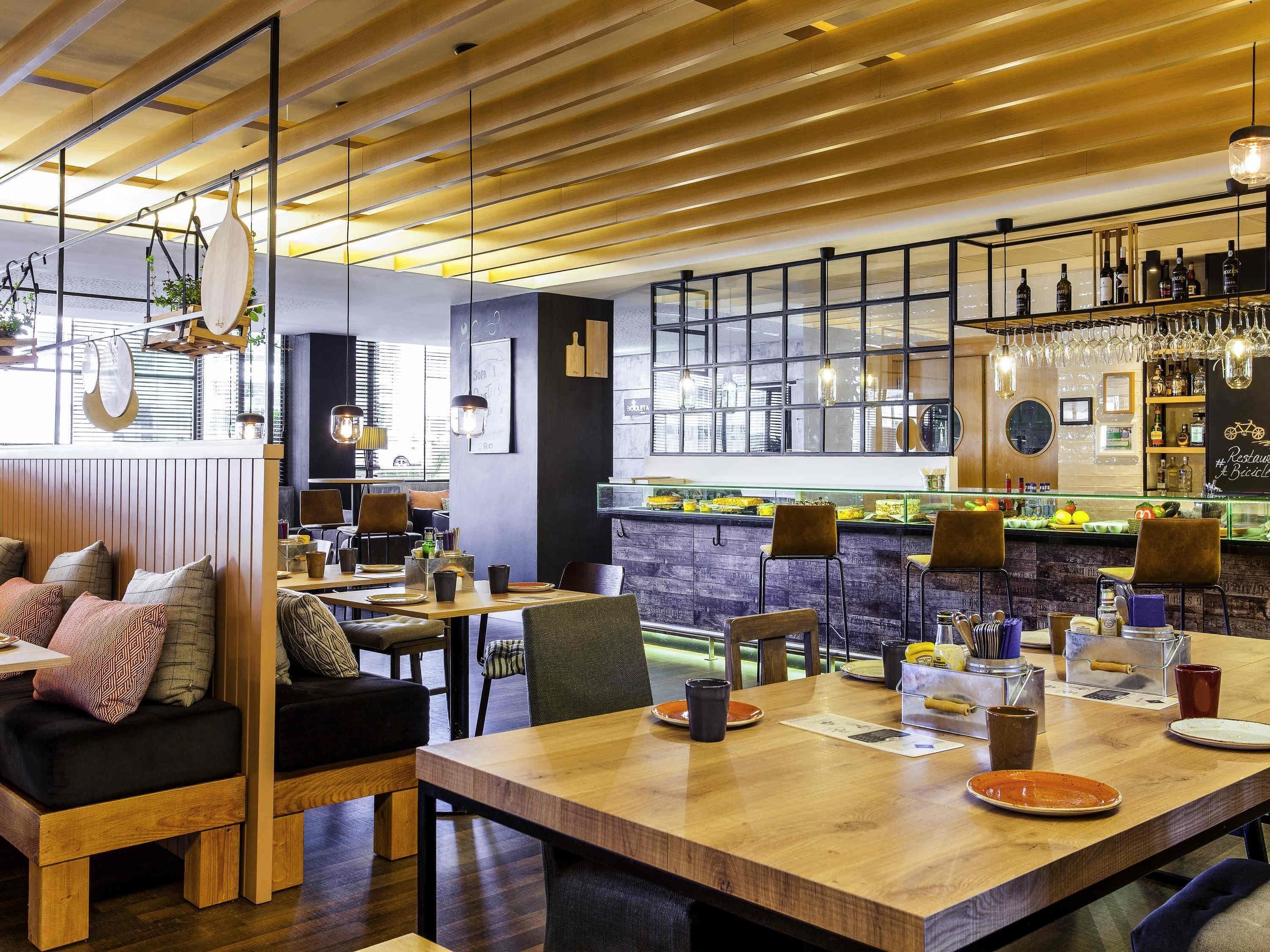Interior moderno de restaurante con mesas de madera, asientos de bar y una exhibición de vinos a la vista.