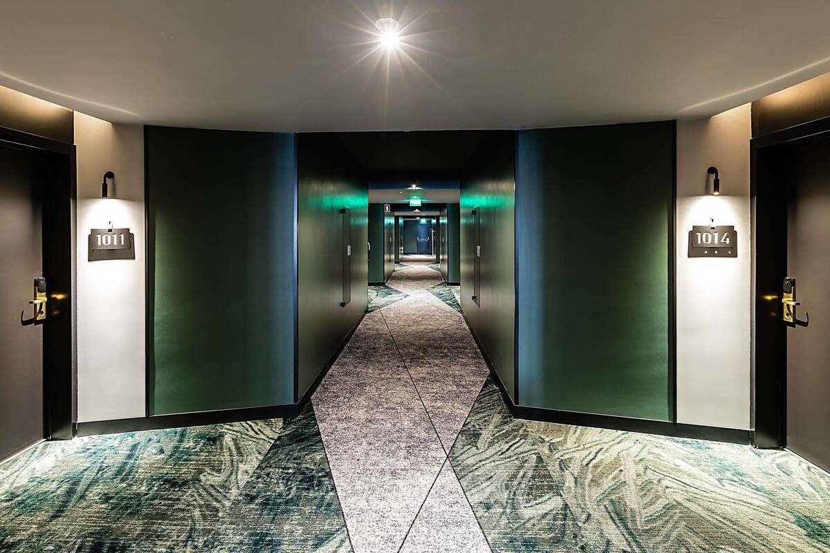 A hotel hallway with a carpeted floor, two doors with illuminated room numbers, and a bright light source at the end.
