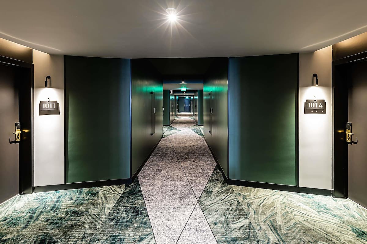 Ein Hotelgang mit Teppichboden, zwei Türen mit beleuchteten Zimmernummern und einer hellen Lichtquelle am Ende.