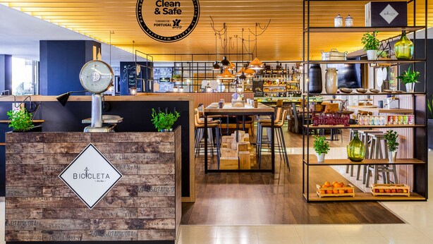 A clean and safe establishment with a wooden reception desk, a dining area with tables and chairs, and shelves with various items. - Novotel Lisboa (Lisbon)
