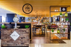 A clean and safe establishment with a wooden reception desk, a dining area with tables and chairs, and shelves with various items. - Novotel Lisboa (Lisbon)