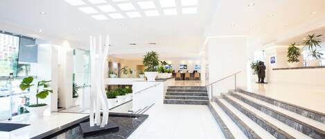 A modern hotel lobby with a marble reception desk, a staircase, and potted plants.