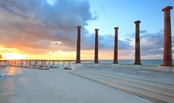 A seaside promenade with steps leading to a beach, featuring columns and a sunset view.