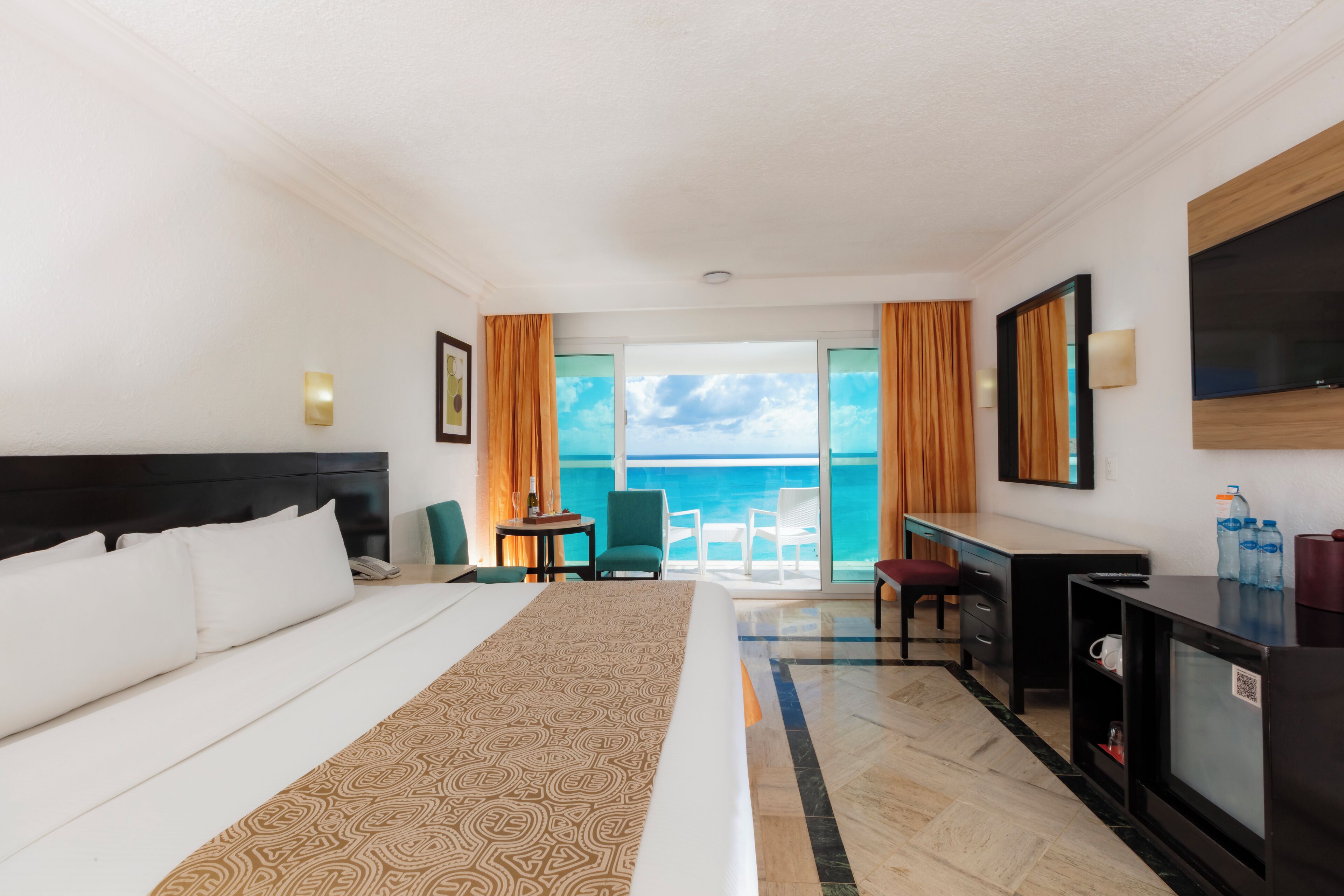 A hotel room with a large bed, a desk, a chair, a TV, and a view of the ocean.