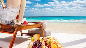 A beachside dining setup with a wooden chair, a tray with food and a bottle of champagne, and a view of the ocean.