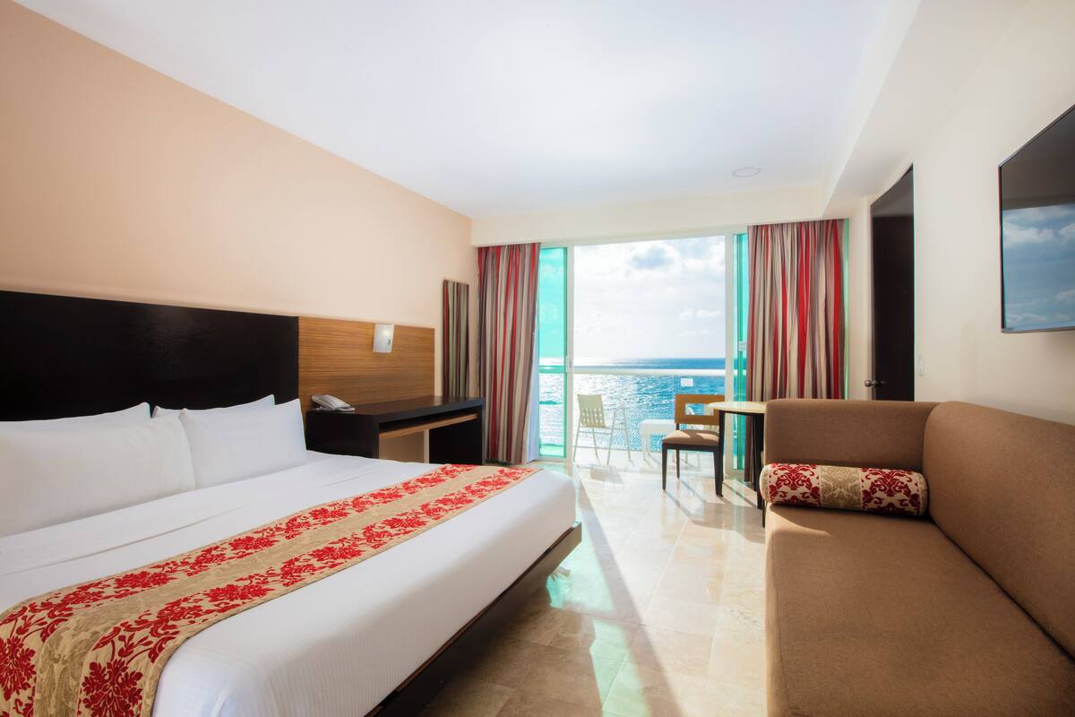 Habitación de hotel con una cama grande, un sofá, un escritorio y un balcón con vistas al mar.