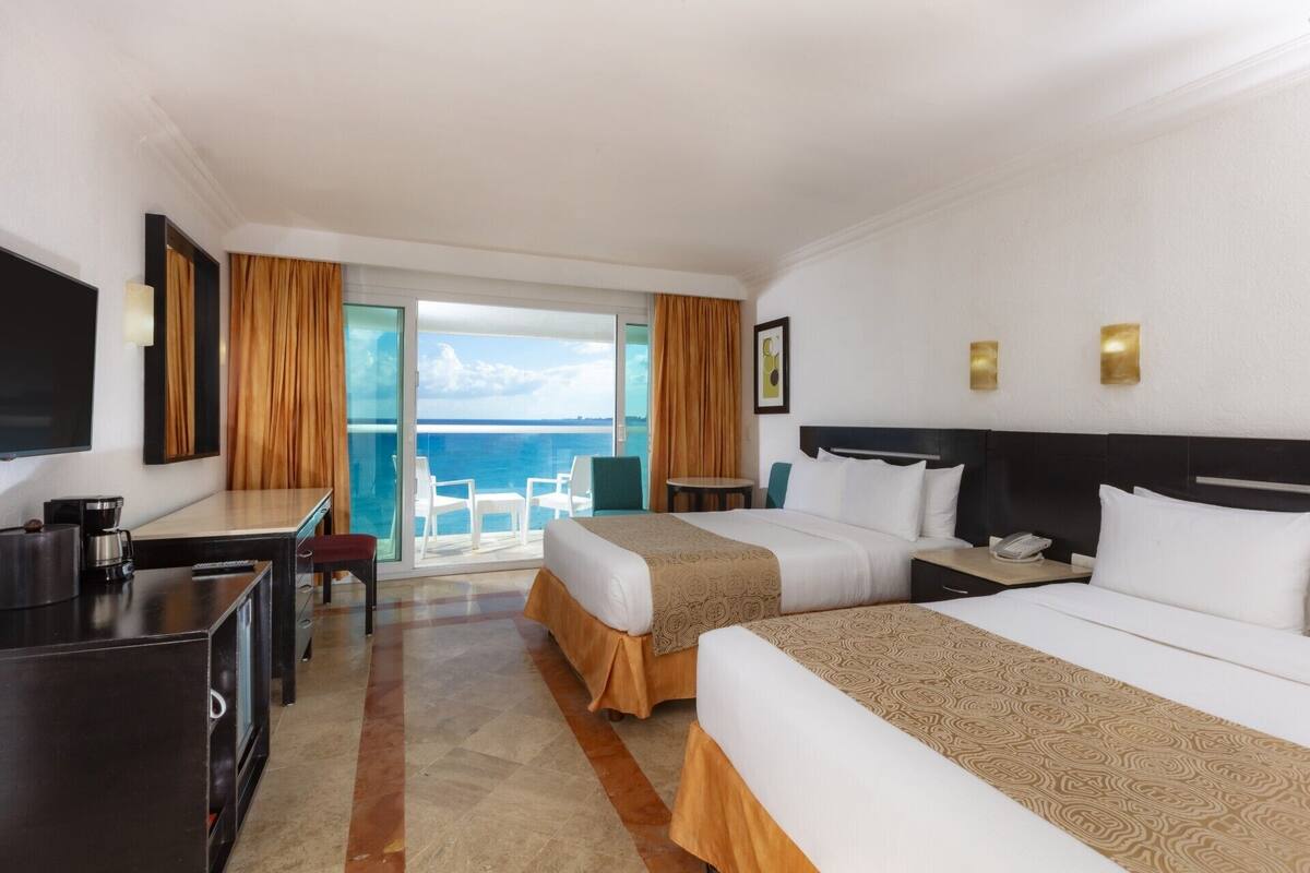 Habitación de hotel con dos camas, un escritorio, una silla, un televisor y un balcón con vista al mar.
