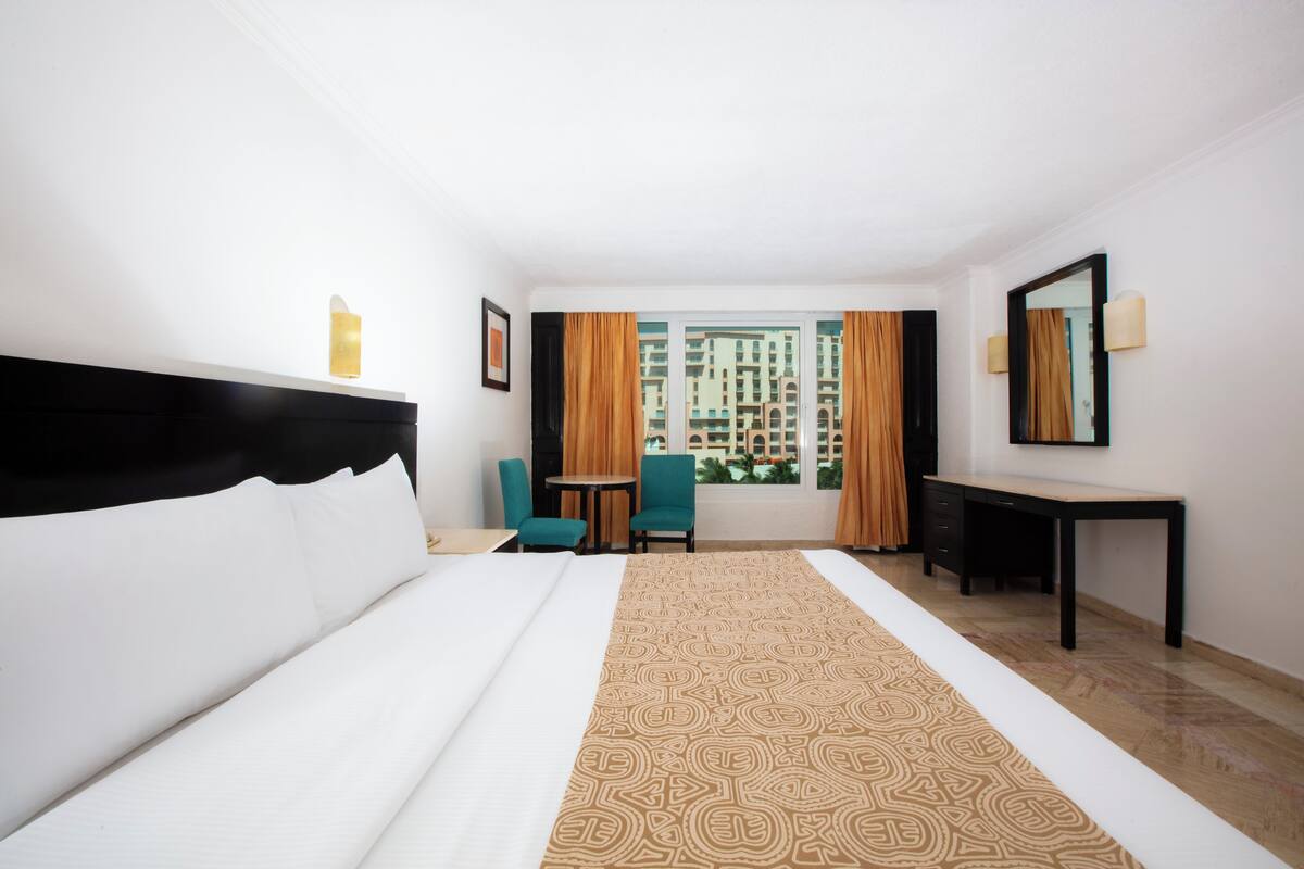 Habitación de hotel con una cama grande, un escritorio, una silla y vistas a edificios a través de la ventana.