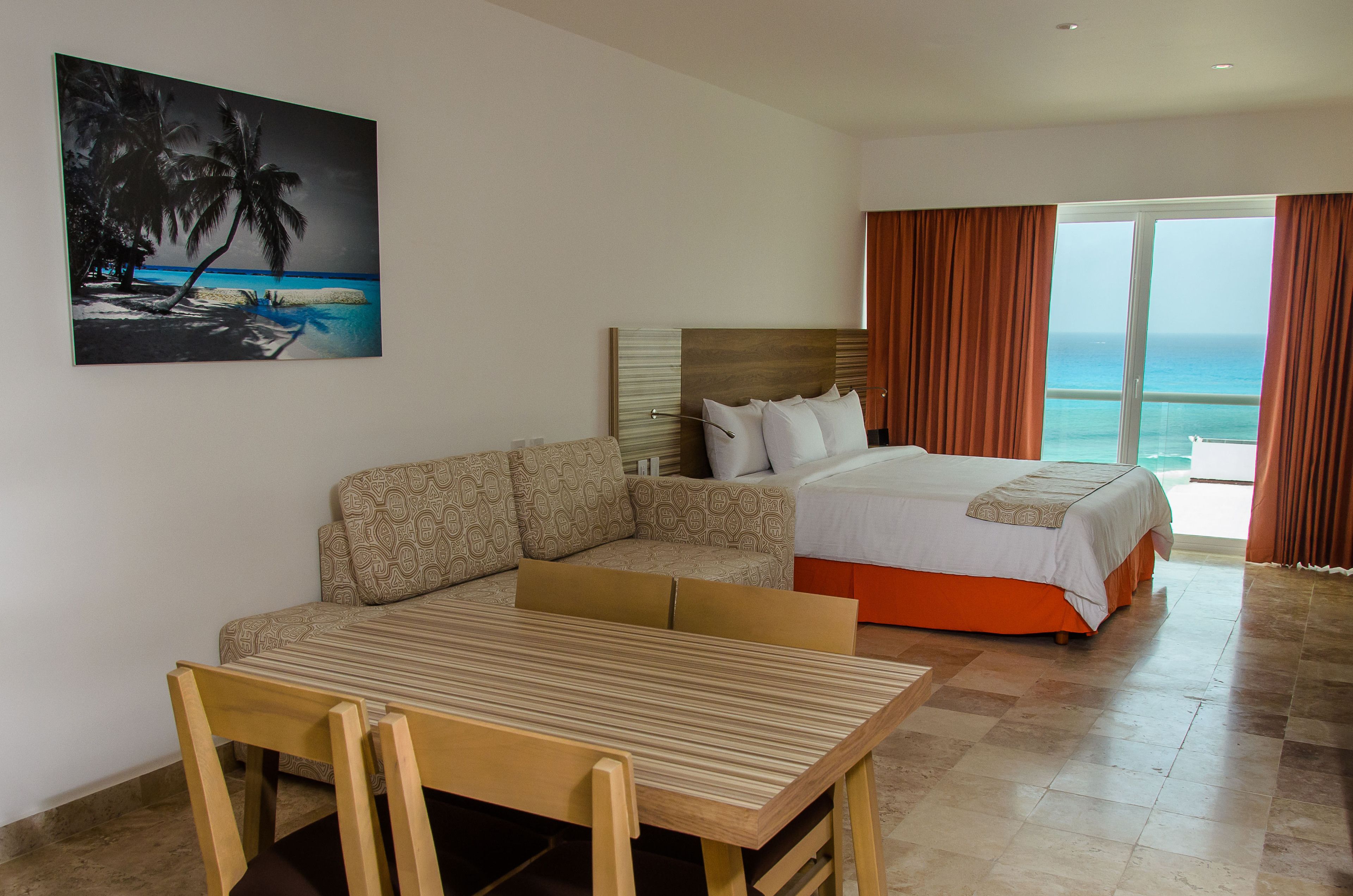 Een hotelkamer met een bed, een bank, een eettafel en een schilderij van een strand.