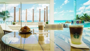 A glass table with a croissant and a Starbucks drink, overlooking a beach with palm trees and a pool.