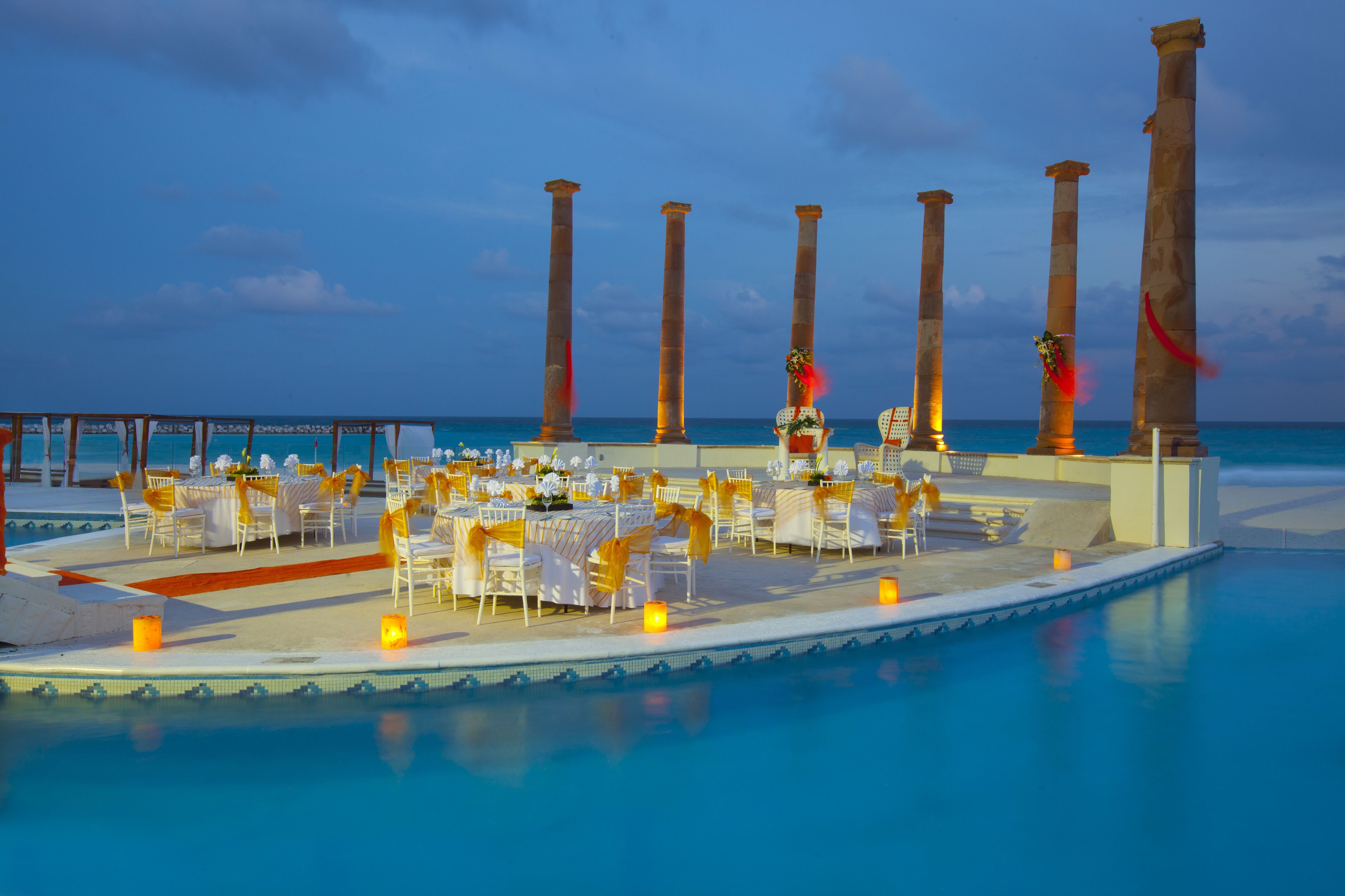 Un área de comedor junto a la piscina con mesas y sillas, columnas y vistas al océano.