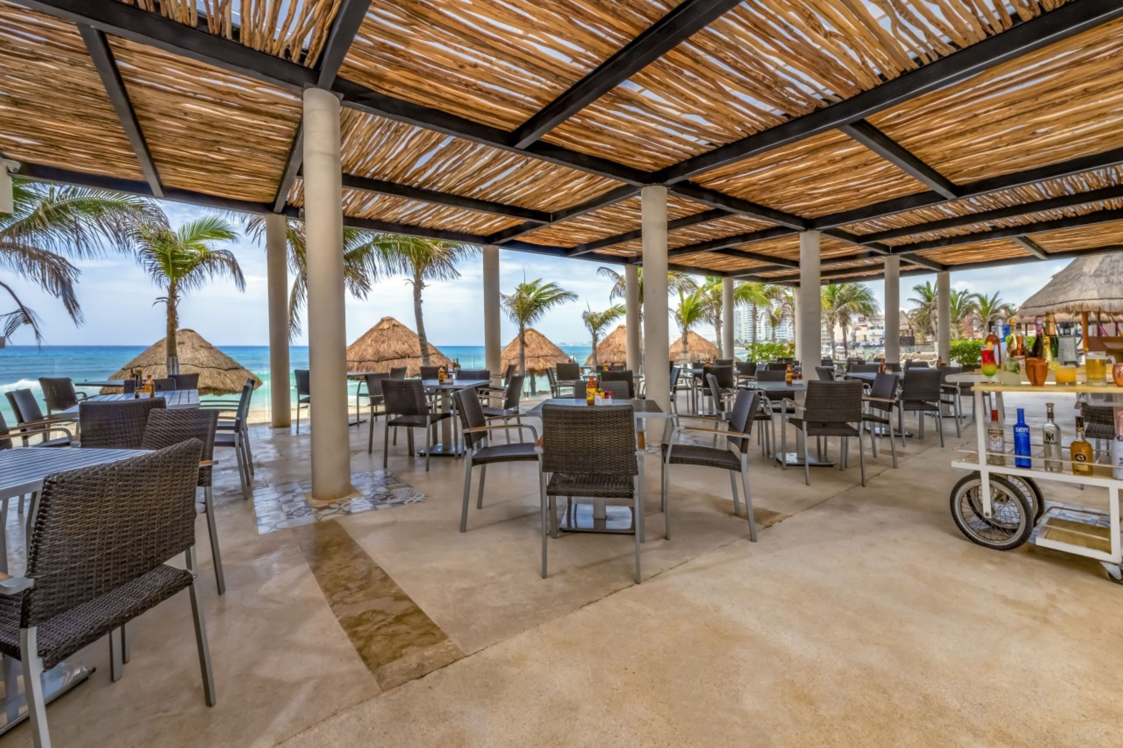 Un espace extérieur aménagé avec des tables et des chaises, un toit de chaume, des palmiers et une vue sur l’océan.