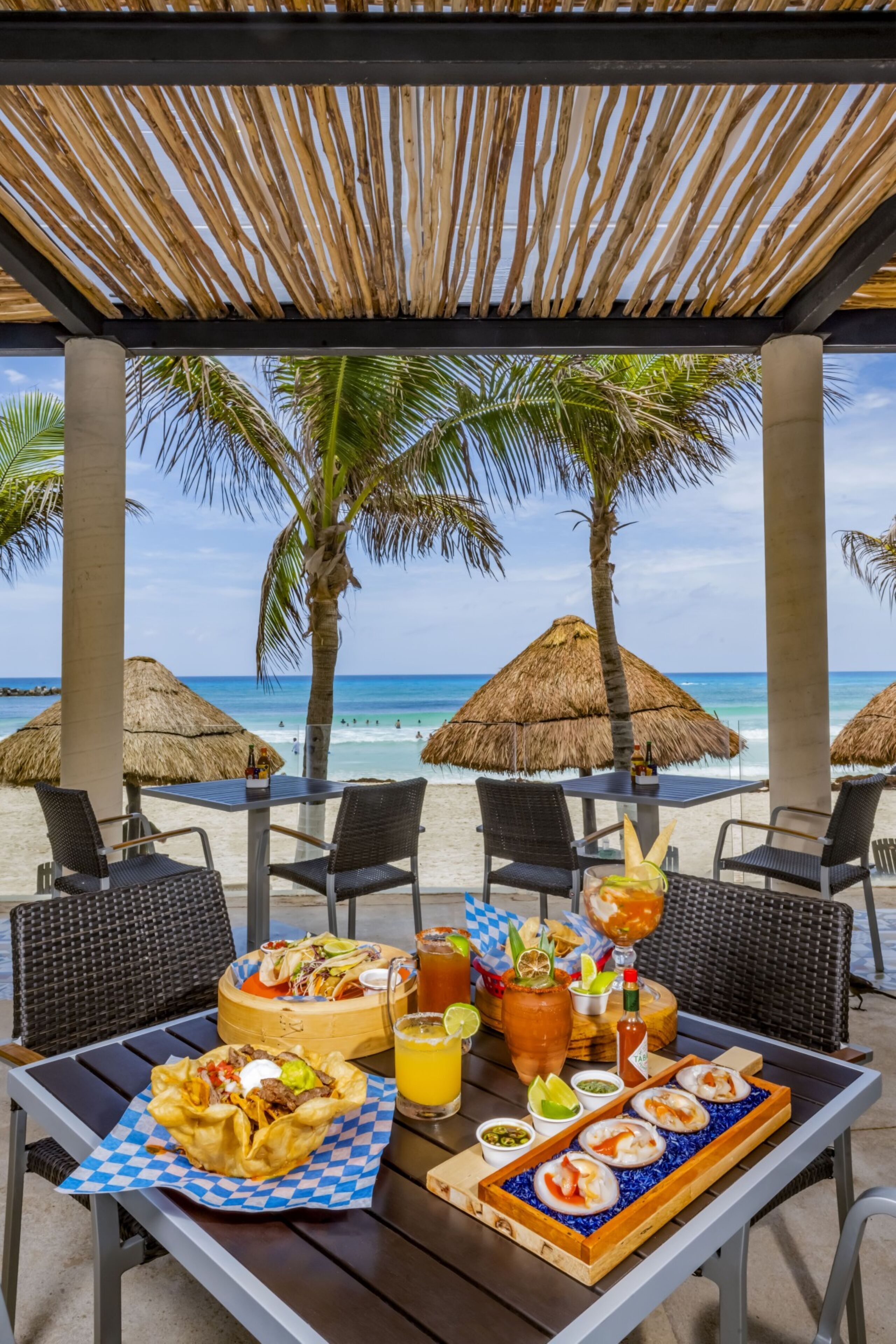 Un área de comedor frente a la playa, con mesas, sillas y sombrillas de paja.