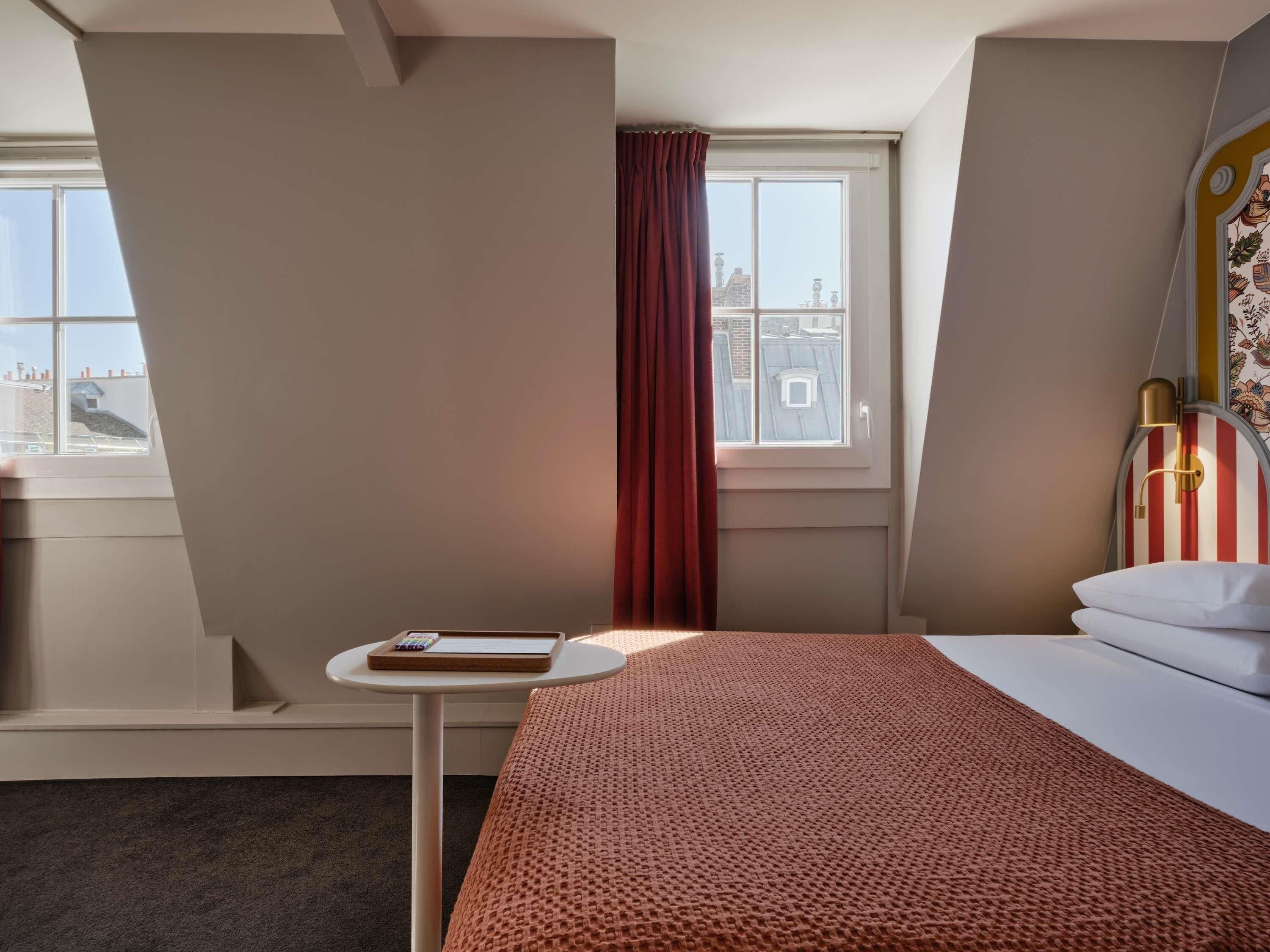 A hotel room with a bed, a small table with a book, a window with a view of buildings, and a curtain.