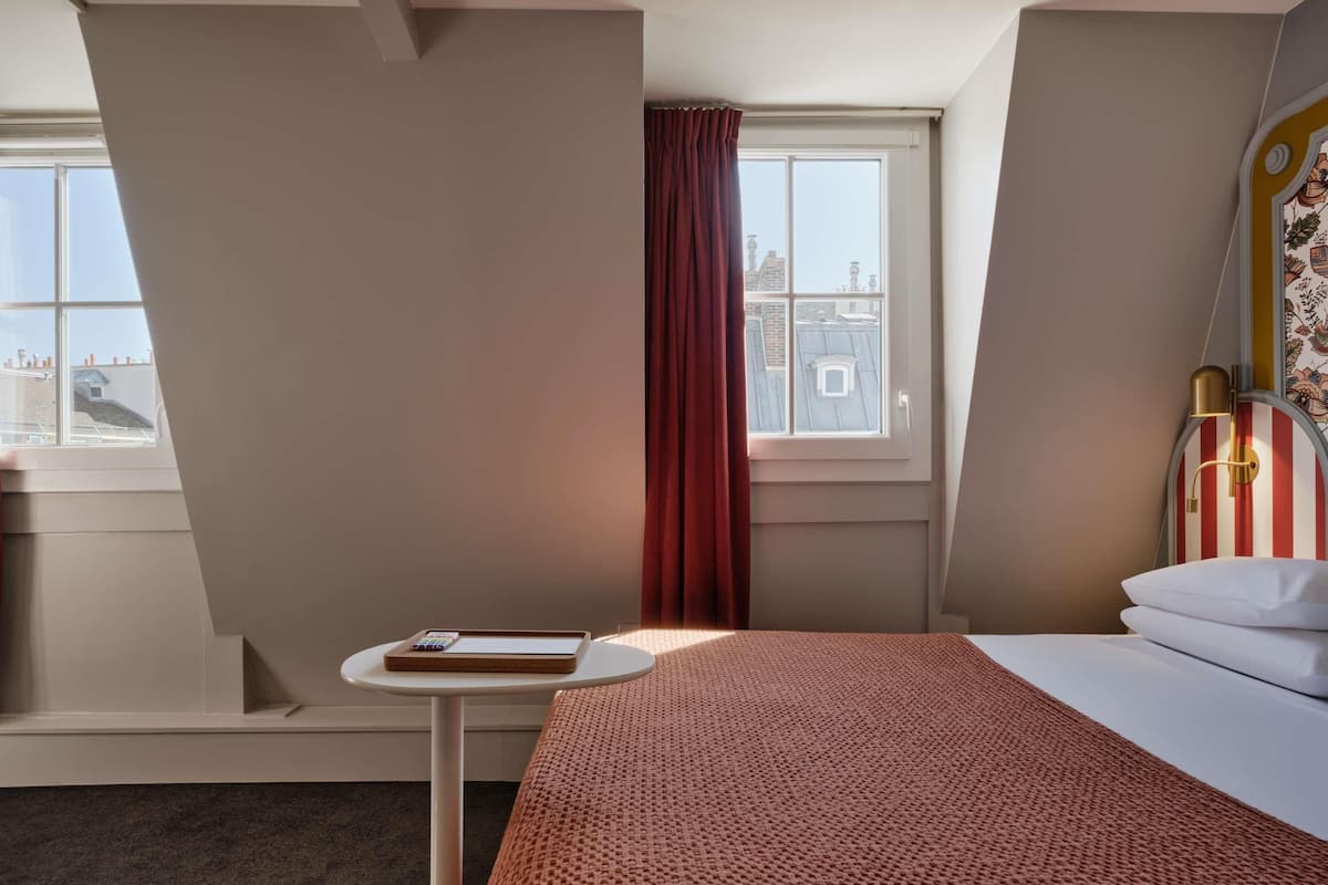 Habitación de hotel con cama, una mesita con un libro, ventana con vistas a edificios y cortina.