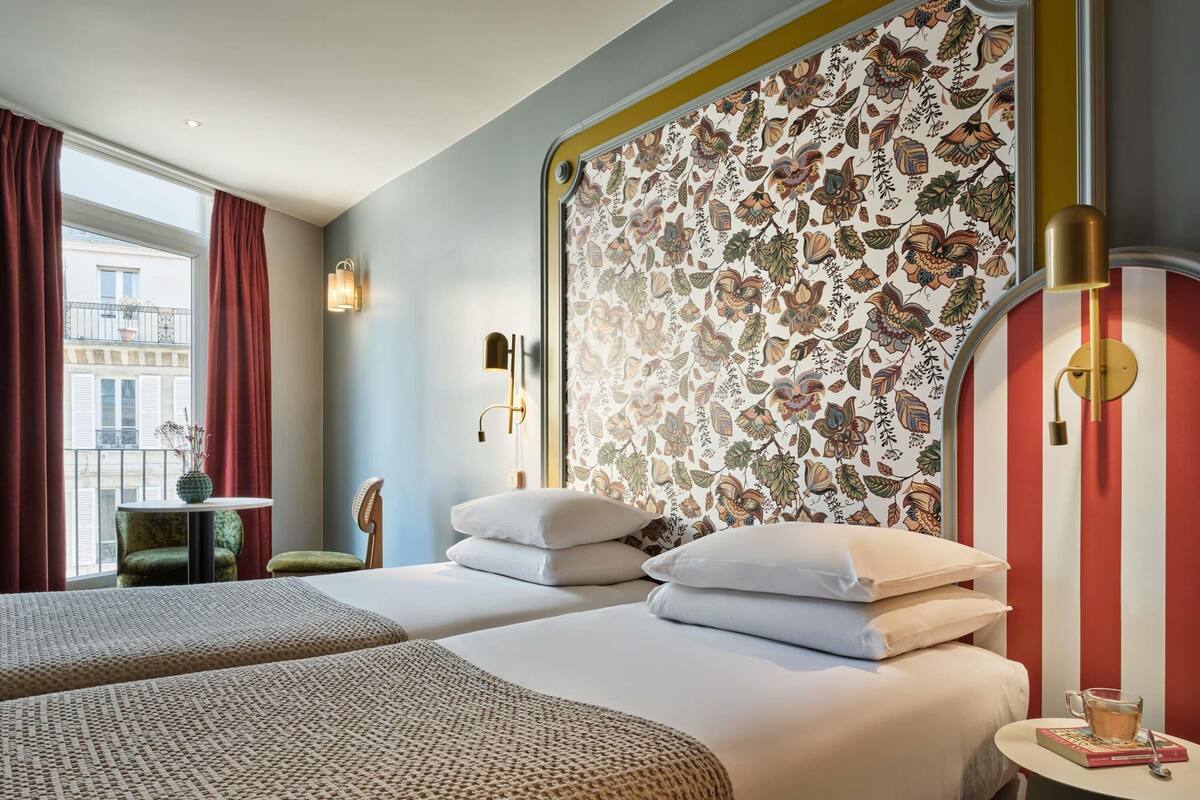 Une chambre d’hôtel avec deux lits, une petite table, une chaise et une fenêtre avec des rideaux.
